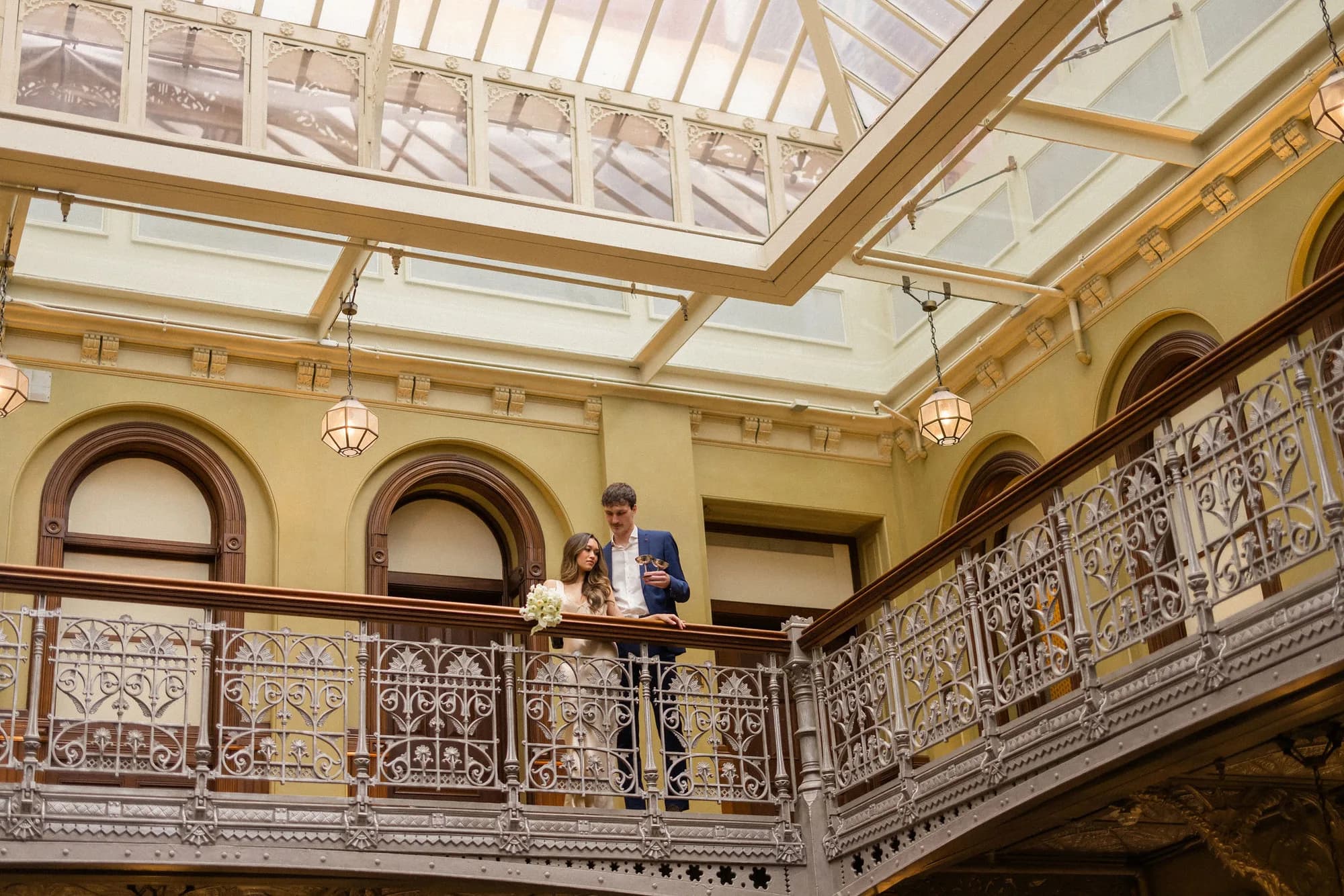 A couple stands at an ornate cast-iron balcony beneath a glass atrium, a white bouquet resting on the wooden rail