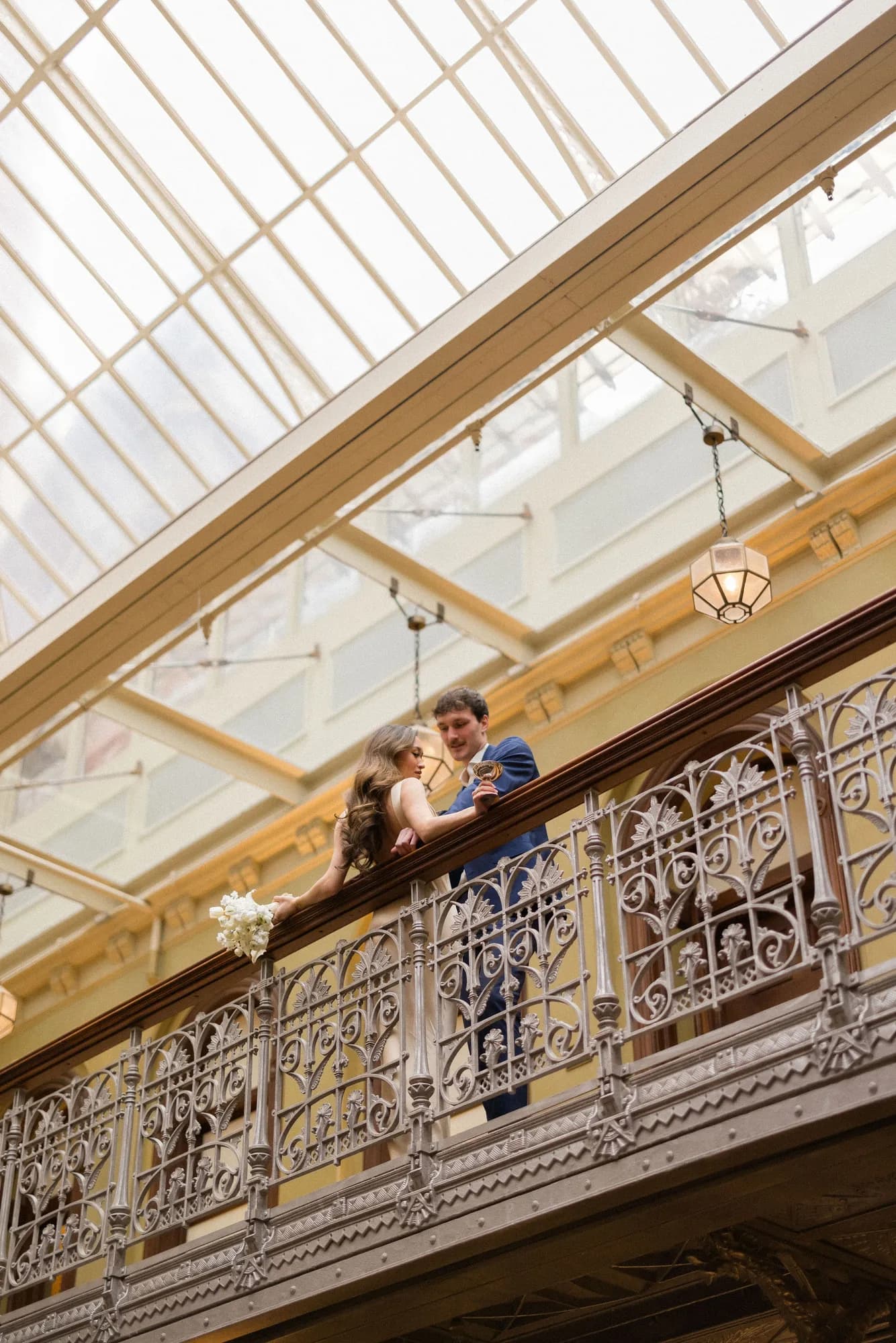 Seen from below, a bride leans on a cast-iron balcony rail with a white bouquet as her groom lifts a coupe under skylights