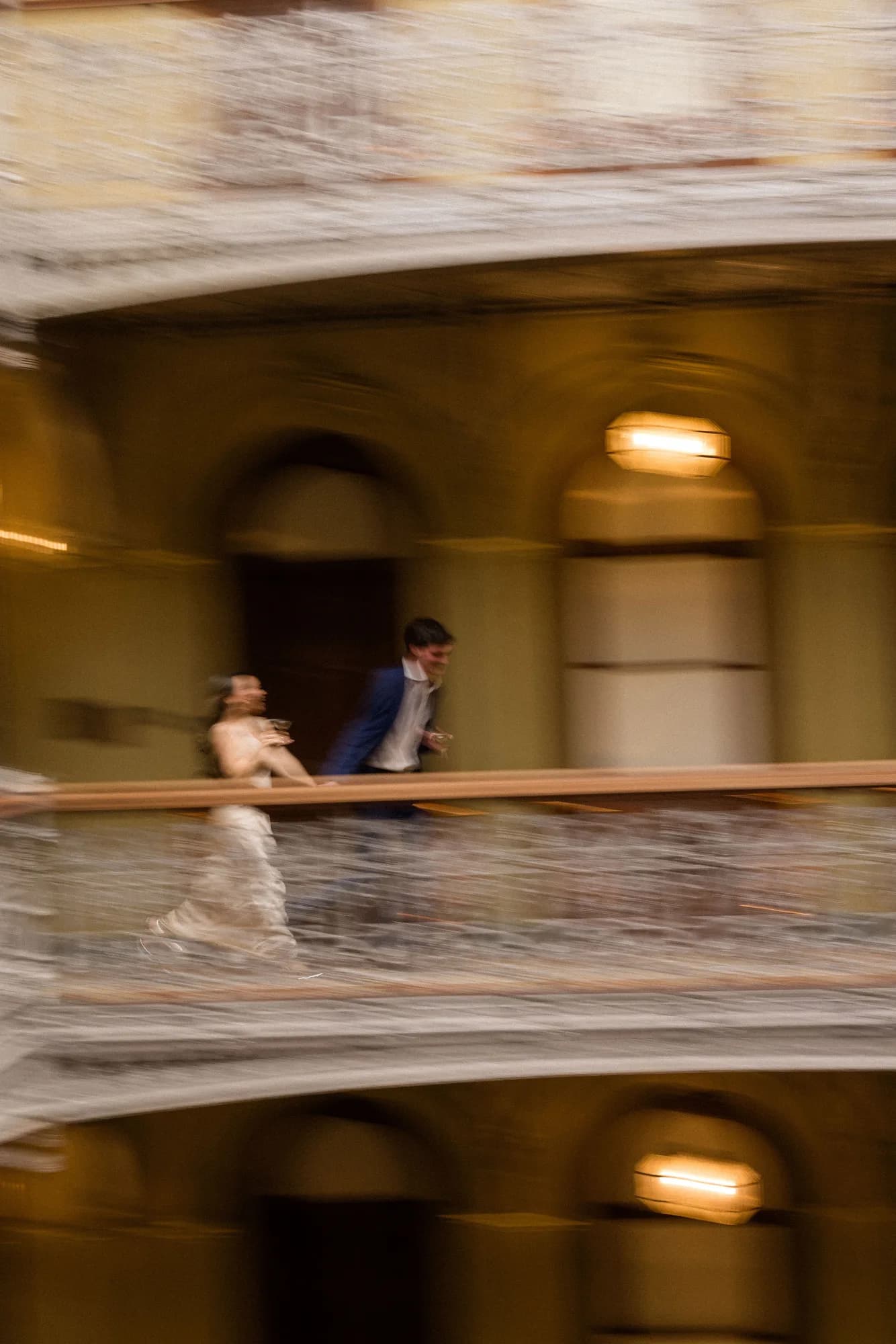 A motion-blurred couple crosses an upper gallery with drinks in hand, a lantern glowing against arched walls