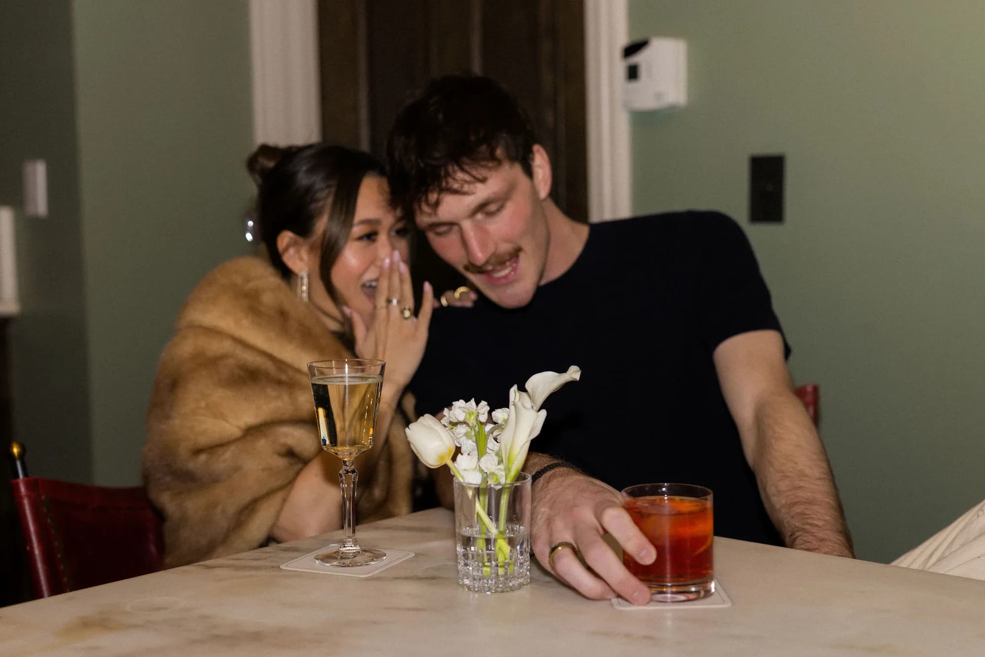 A couple leans together at a marble bar over a negroni and a coupe, white tulips and a calla lily in a cut crystal glass