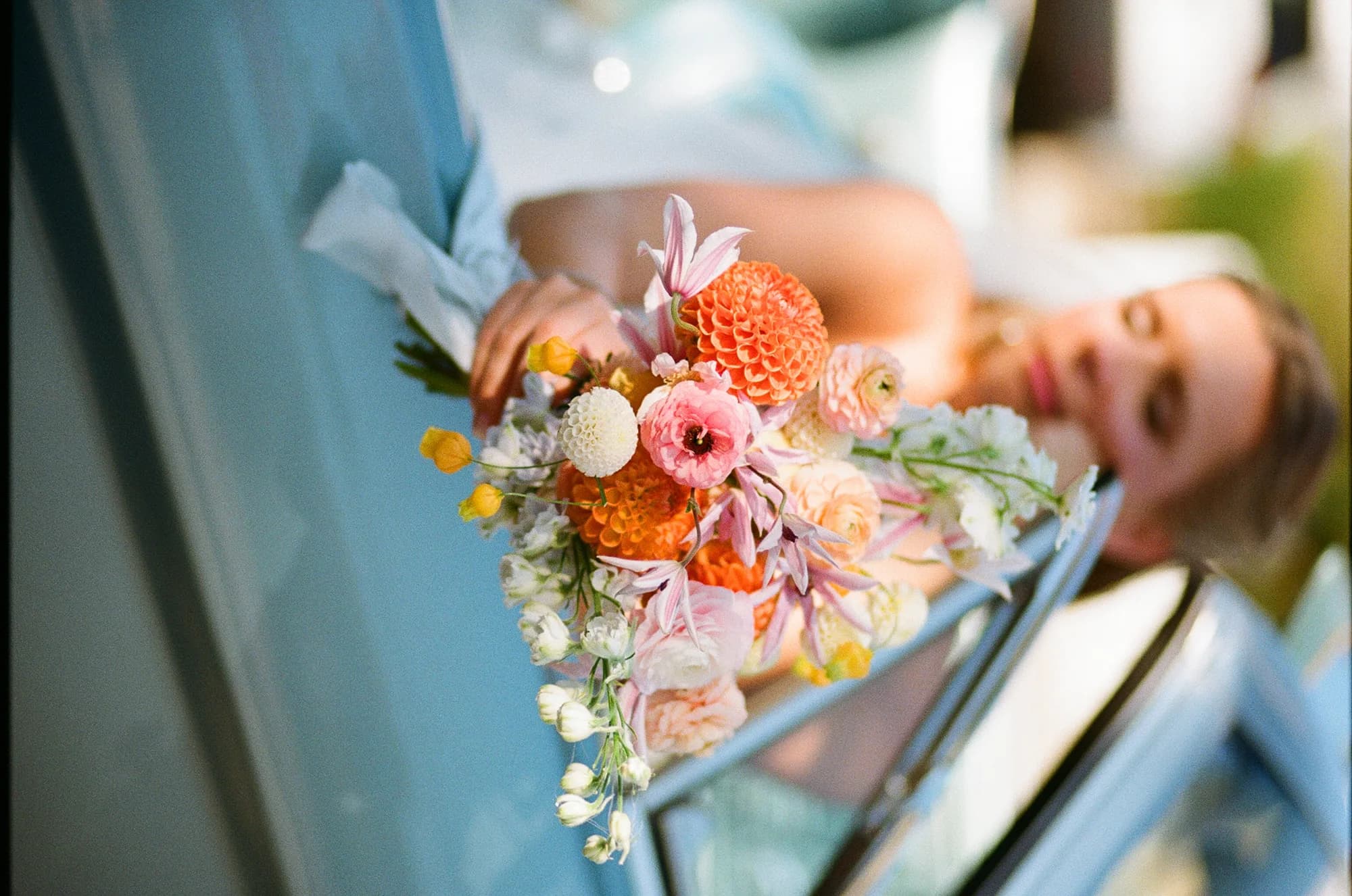 Talea leans from a pale blue convertible window holding a bouquet of orange dahlia, clematis and sandersonia