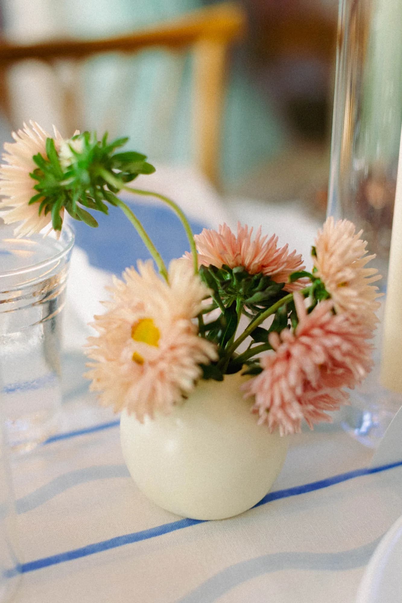 Peach aster stems arc from a small white bud vase on blue-striped linen beside a water glass