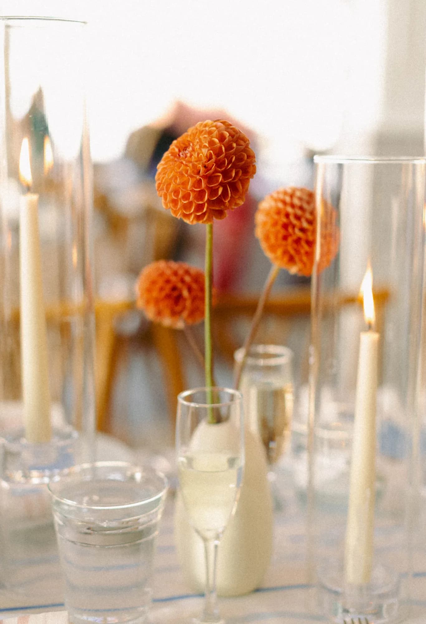 Three orange ball dahlias rise from a white bud vase among tall glass candle sleeves and lit tapers