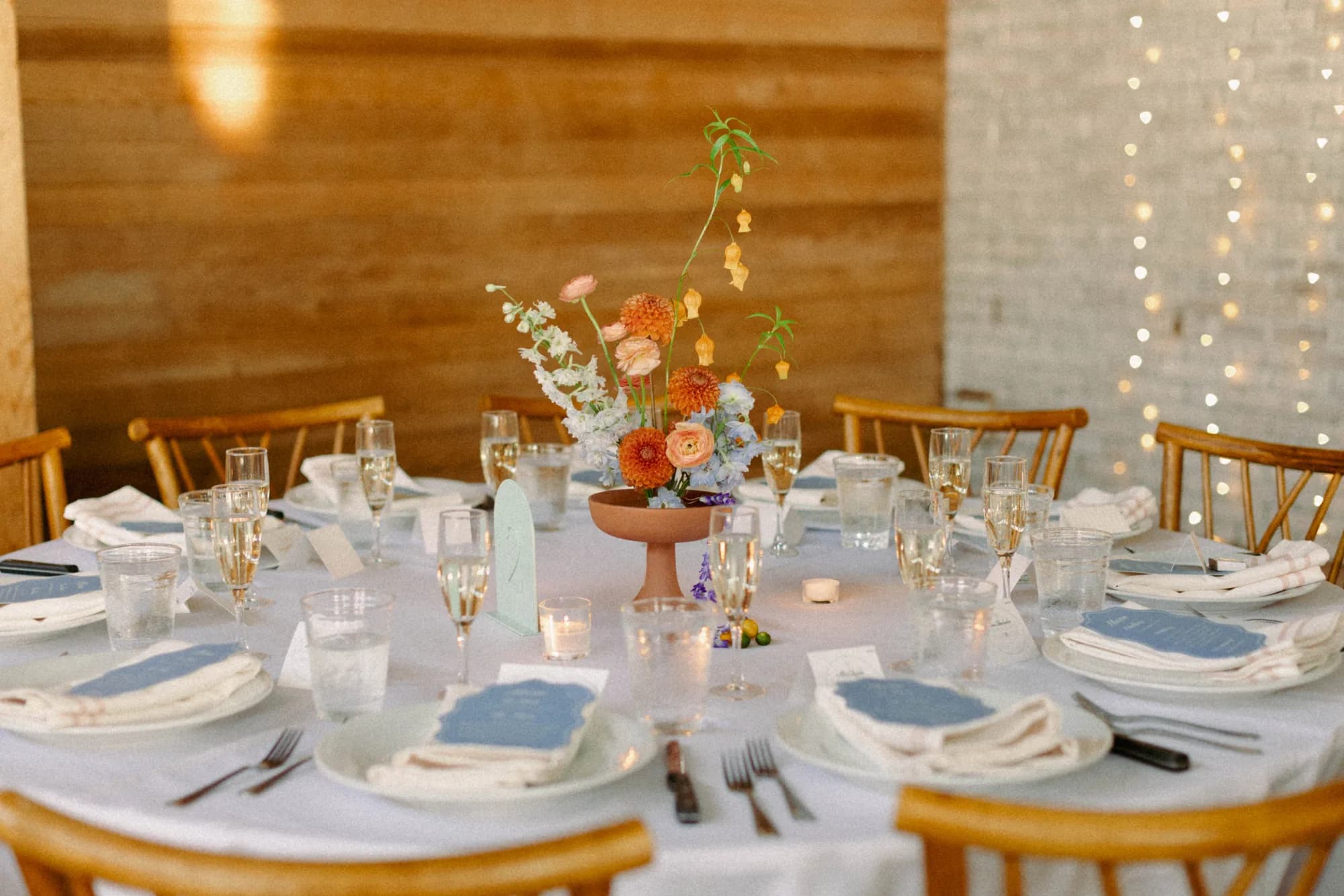 Round reception table with a terracotta compote of orange dahlias, blue delphinium and arching sandersonia