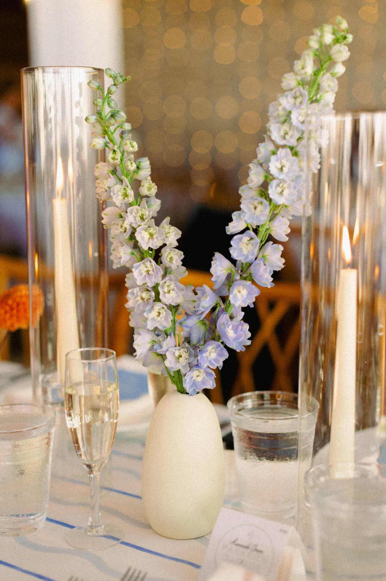 Two pale blue delphinium stems in a white bud vase between glass hurricane sleeves and tapers, lights glowing behind