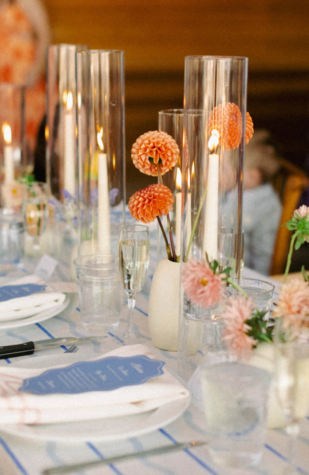 Peach ball dahlias in a white bud vase along a long table lined with glass candle sleeves and blue menu cards