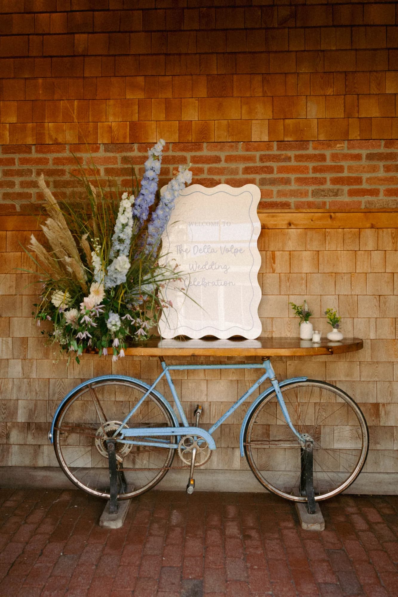 Welcome sign on a wooden table set over a blue vintage bicycle, with blue delphinium, dried grasses and clematis