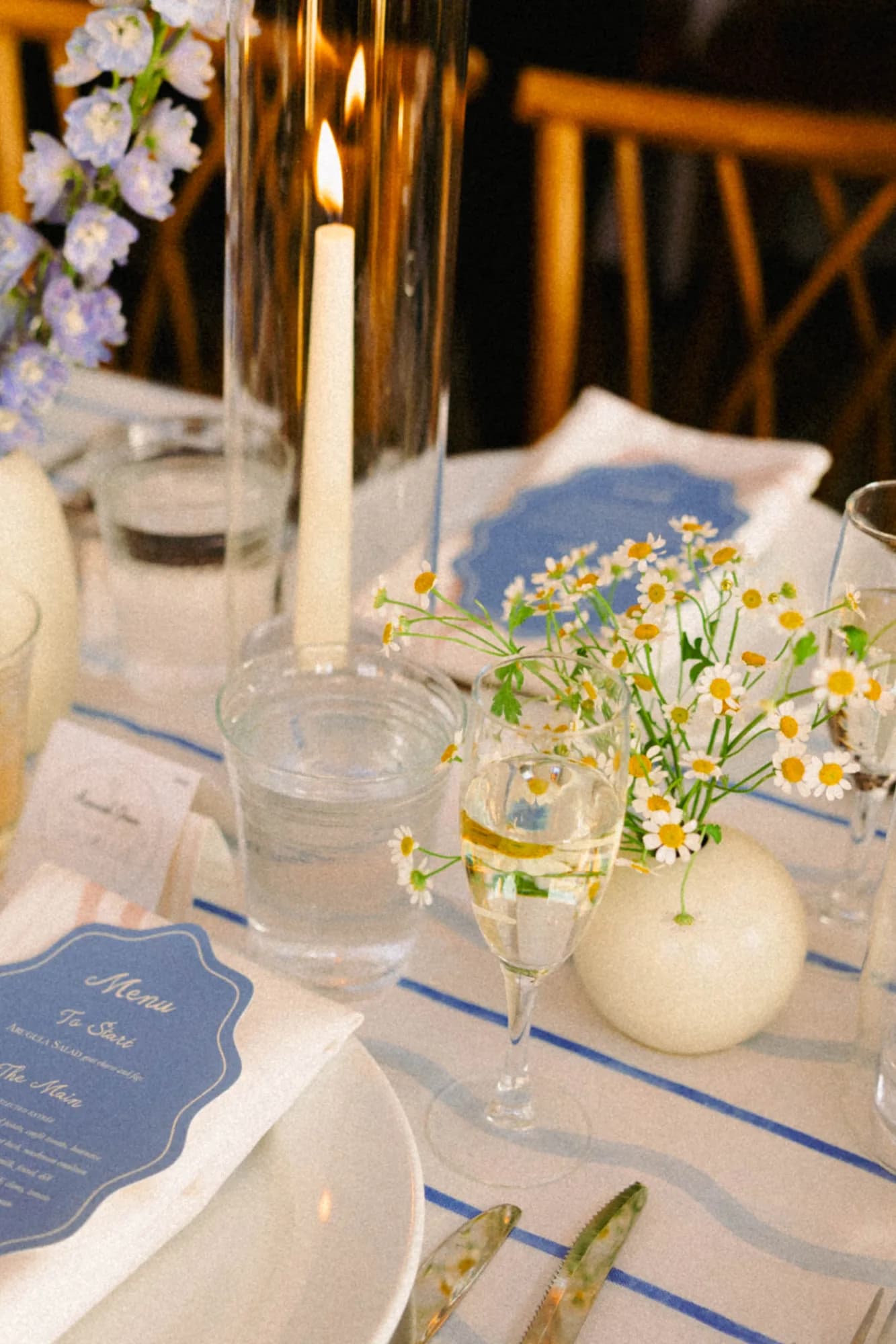 Feverfew daisies in a round white bud vase beside a champagne flute, blue menu card and lit taper on striped linen