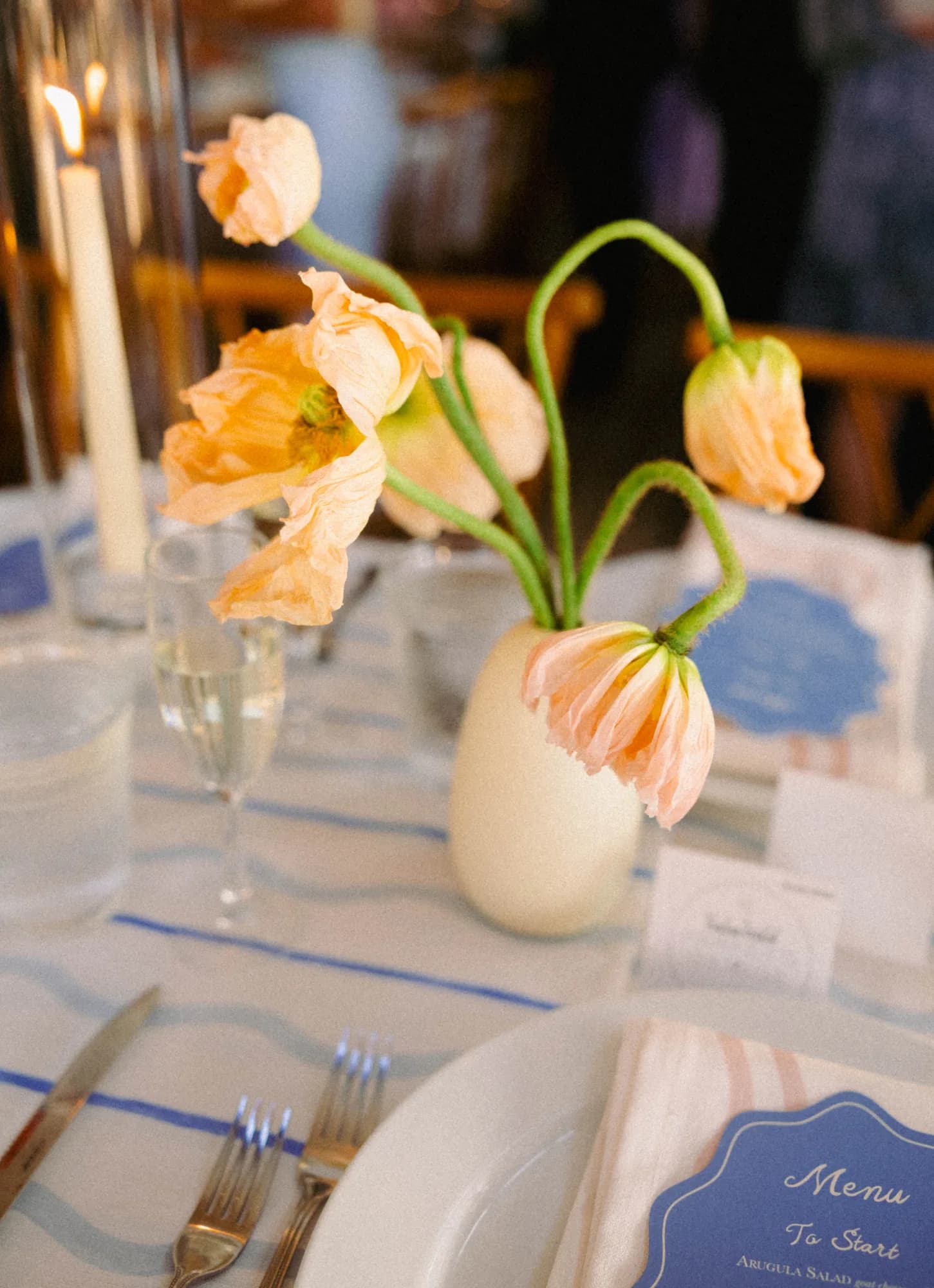 Peach Icelandic poppies on curving stems lean from a white bud vase beside candlelight and a blue menu card
