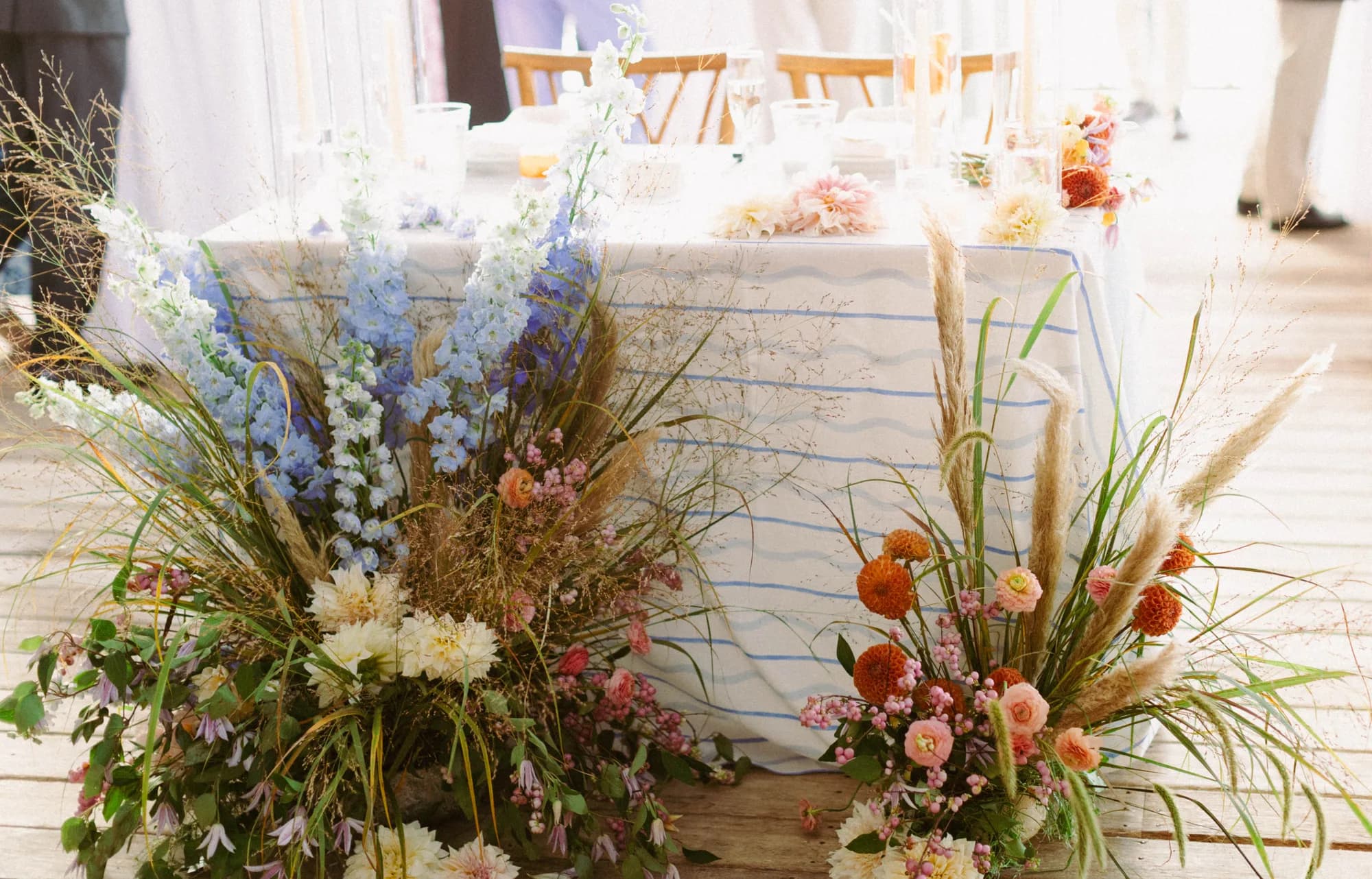 Two ground arrangements of blue delphinium, dried grasses, orange dahlias and snowberry flank the head table