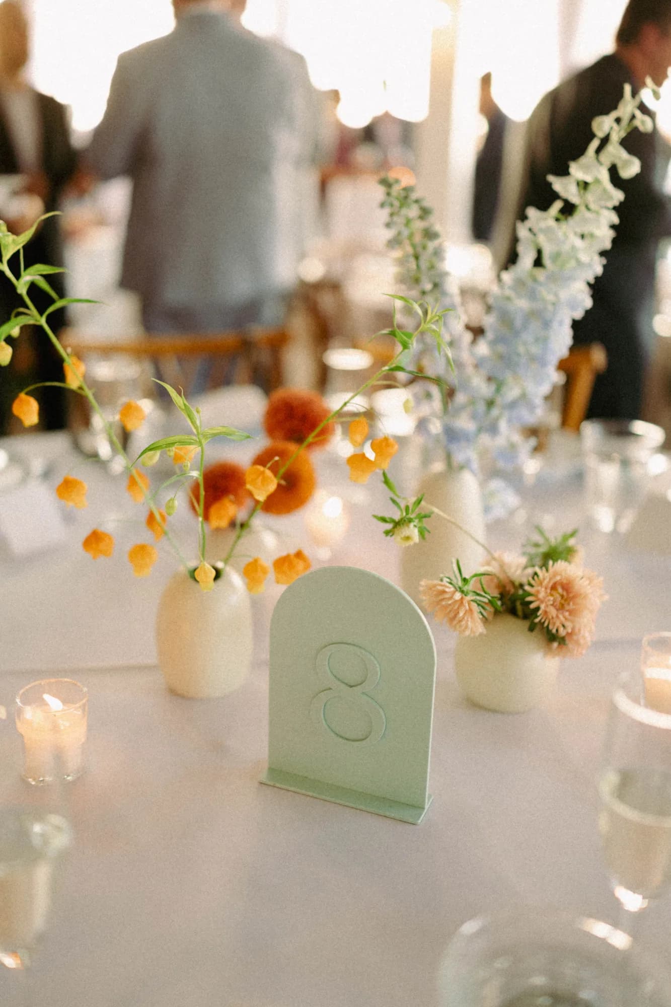 Mint table number eight among bud vases of sandersonia, pale blue delphinium and peach asters, orange dahlias behind