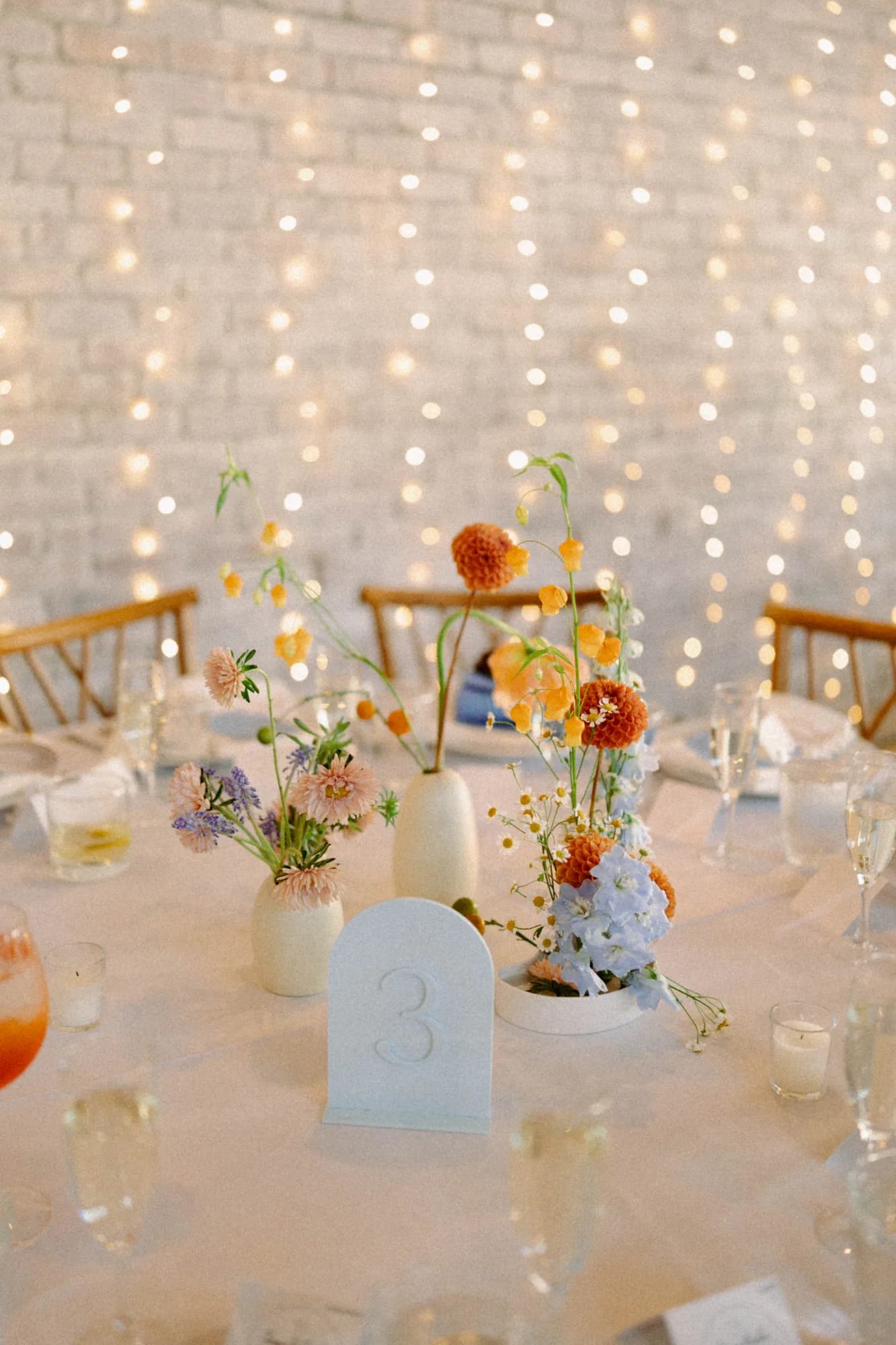 Table number three with bud vases of peach asters, orange dahlias and sandersonia against a wall of string lights