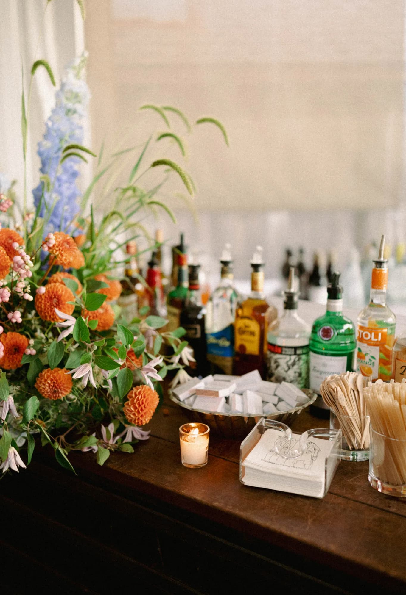 Orange dahlias, blue delphinium, snowberry and clematis spill from a bar arrangement beside rows of bottles