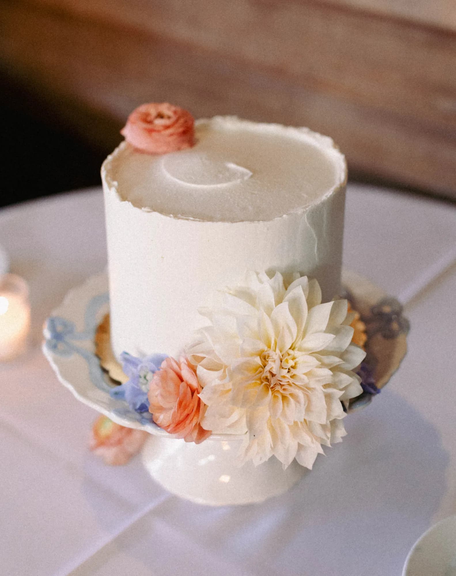 Single-tier white cake topped with a peach ranunculus, a cream dahlia and blue delphinium tucked at its base