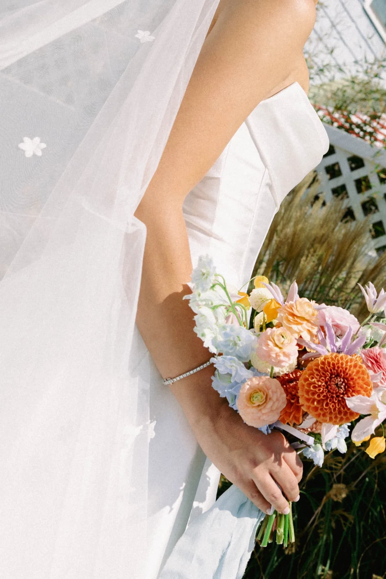 Talea holds her bouquet at her hip, orange dahlia, peach ranunculus and blue delphinium against a lattice fence