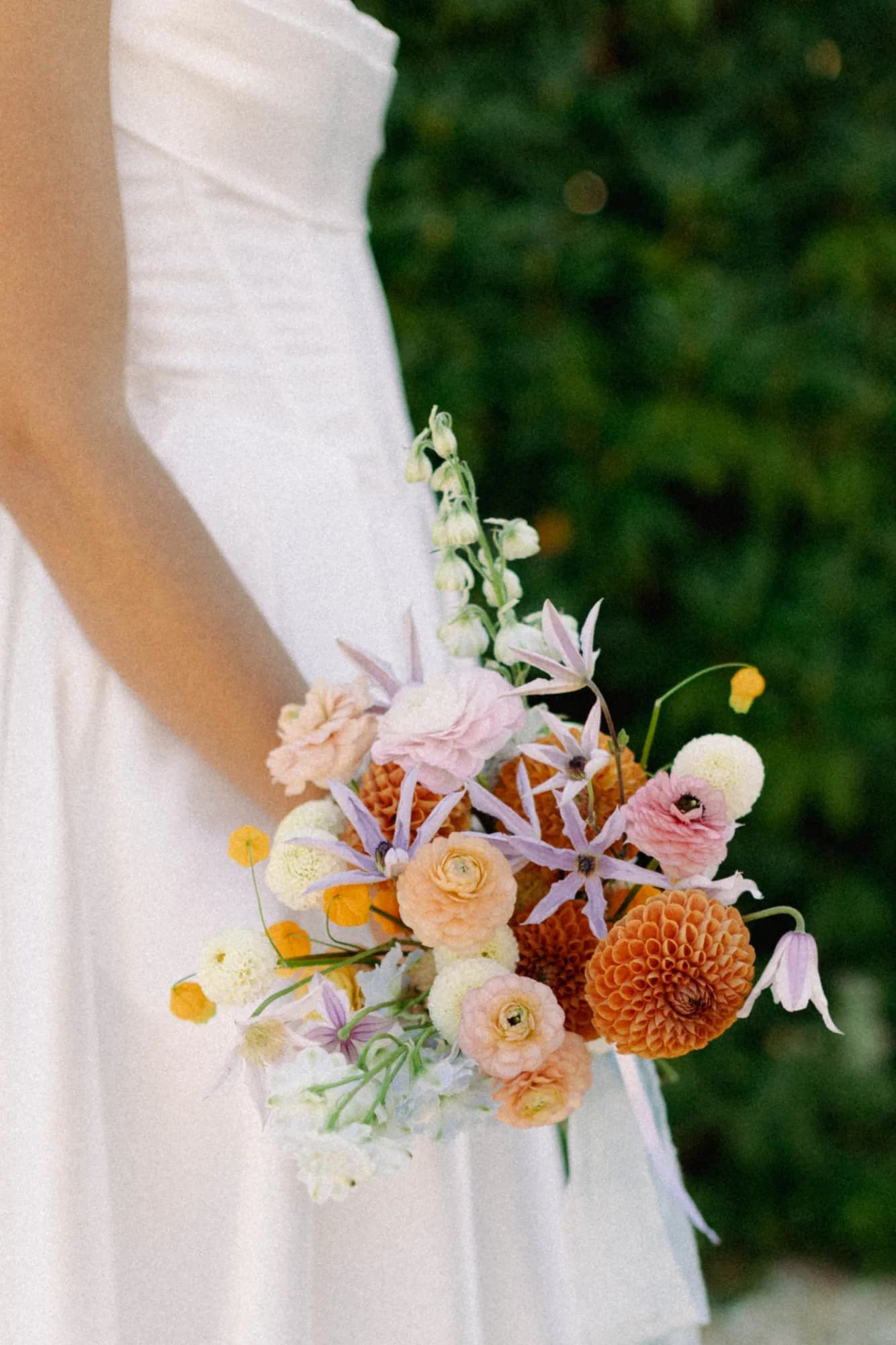 Talea holds a bouquet of orange dahlias, peach ranunculus, lavender clematis and sandersonia against a green hedge