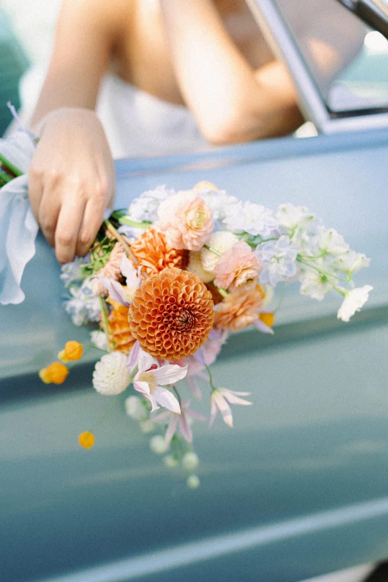 Talea rests her bouquet on the door of a pale blue convertible, orange dahlia and clematis trailing downward