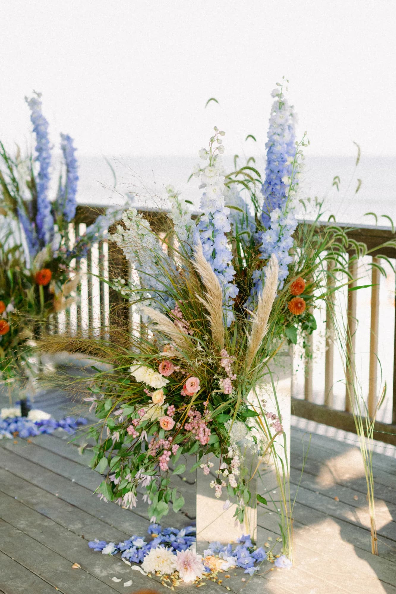 Ceremony arrangement on a mirror plinth with blue delphinium spires, pampas grass and orange dahlias, ocean beyond