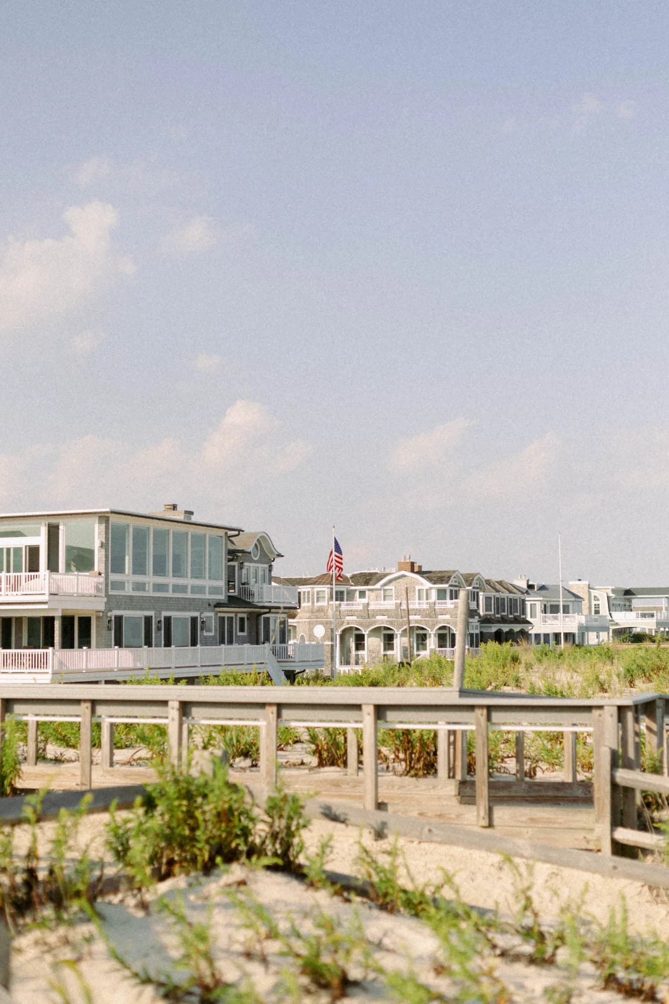 Shingled beach houses and a flag seen across dune fencing and beach grass under a hazy blue sky