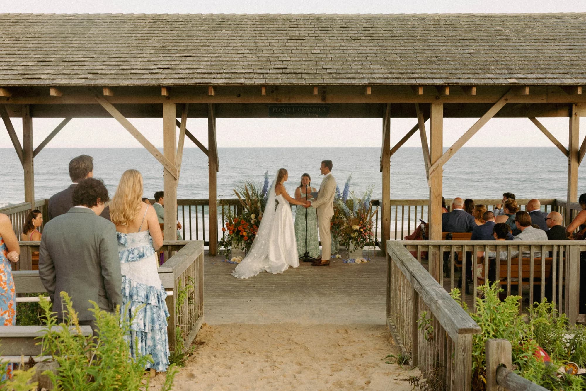 Wide view of the oceanfront pavilion ceremony, Talea and Erich at the altar between two floral arrangements