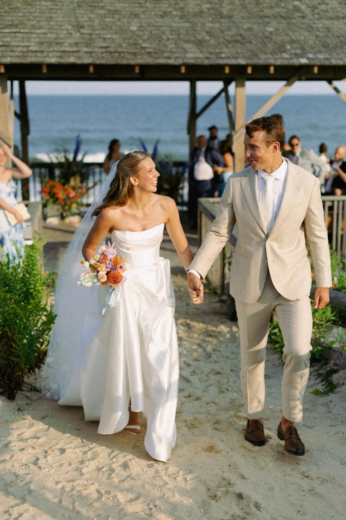 Talea and Erich walk hand in hand up a sandy path from the pavilion, her bouquet trailing pale blue ribbon