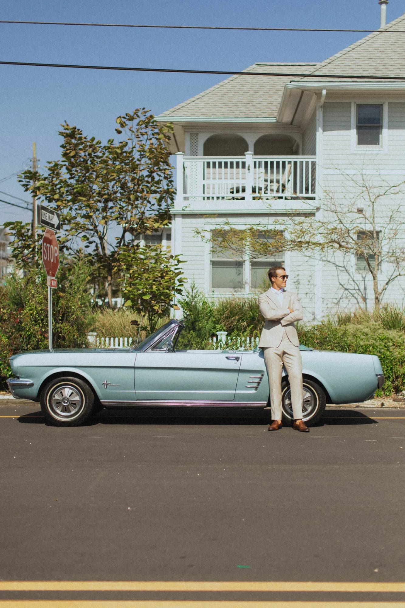 Erich in a cream suit leans against a pale blue vintage convertible in front of a white shingled house