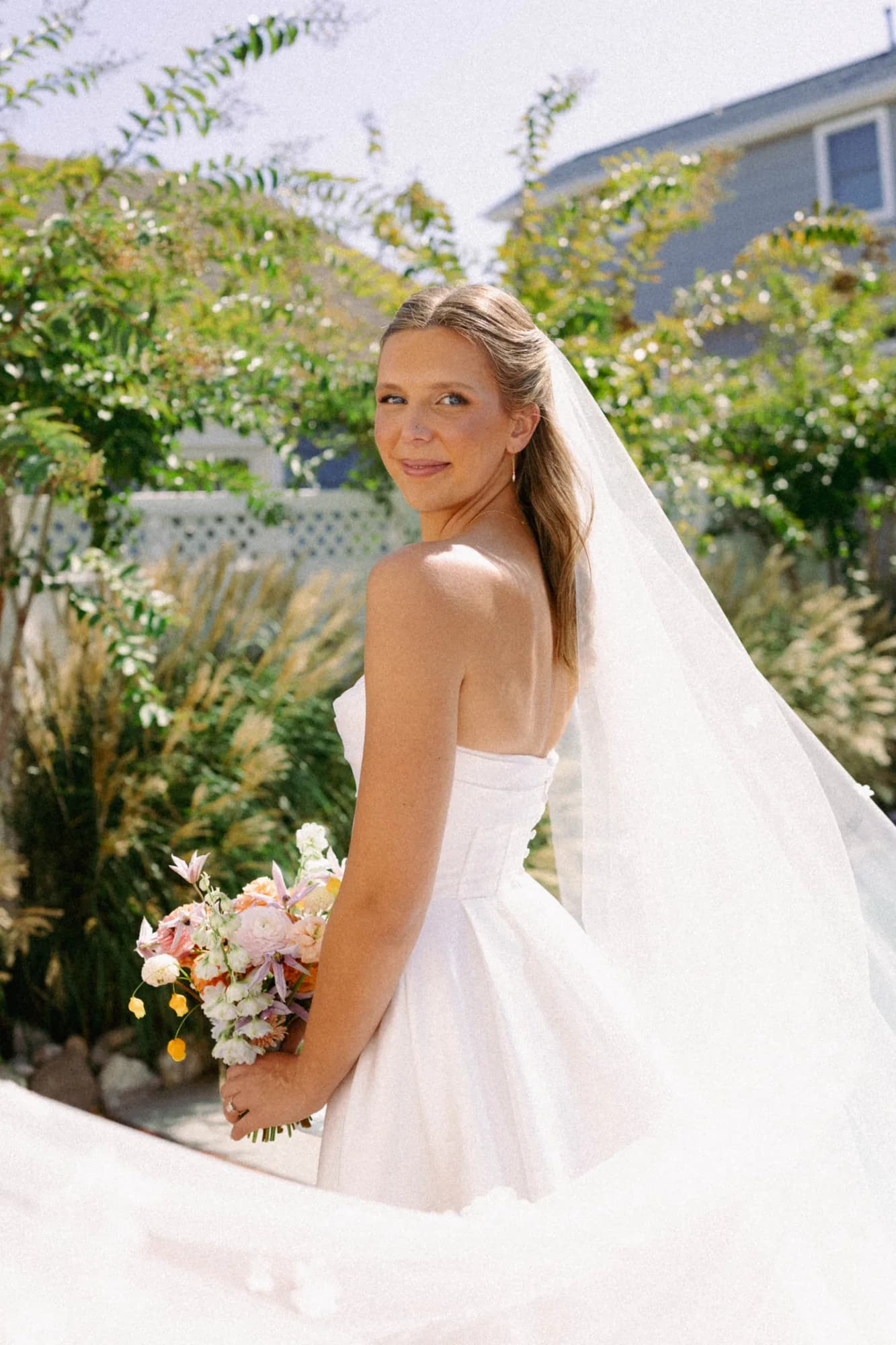 Talea looks back over her shoulder holding a bouquet of clematis, orange dahlias and sandersonia at her hip