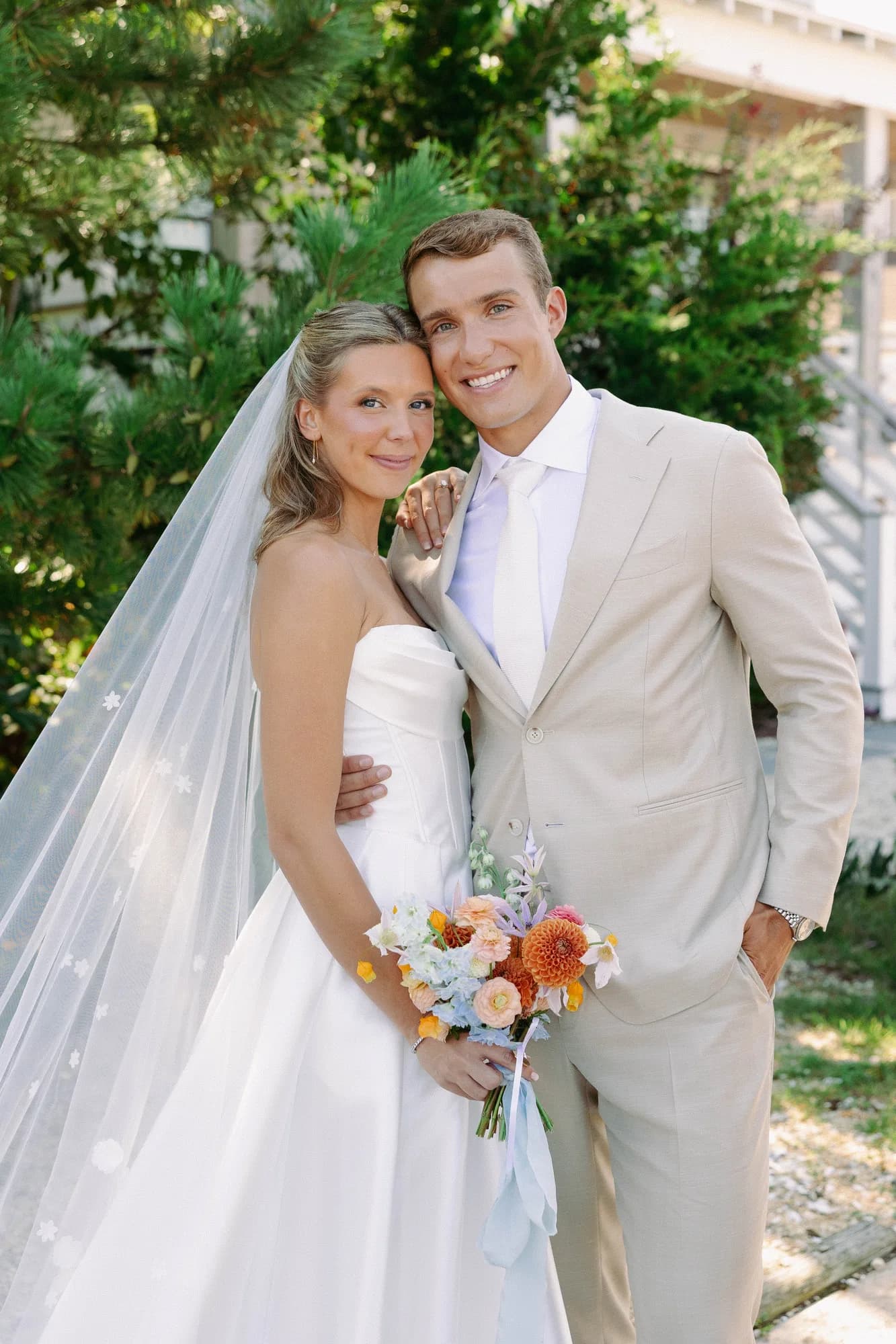 Talea and Erich pose cheek to cheek, her bouquet of orange dahlias, peach ranunculus and blue delphinium in hand