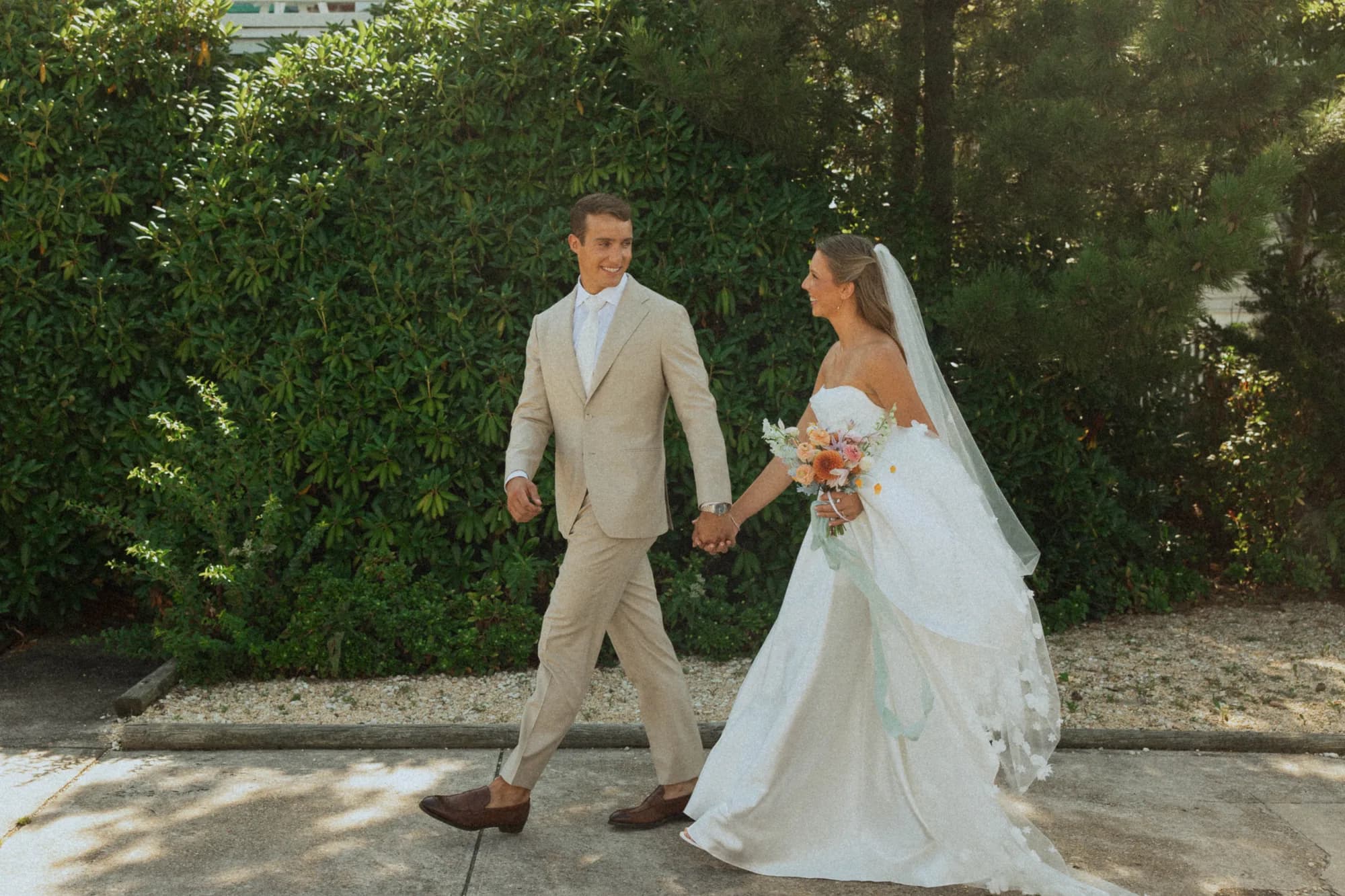 Talea and Erich walk hand in hand along a hedge, her bouquet trailing a long pale blue ribbon