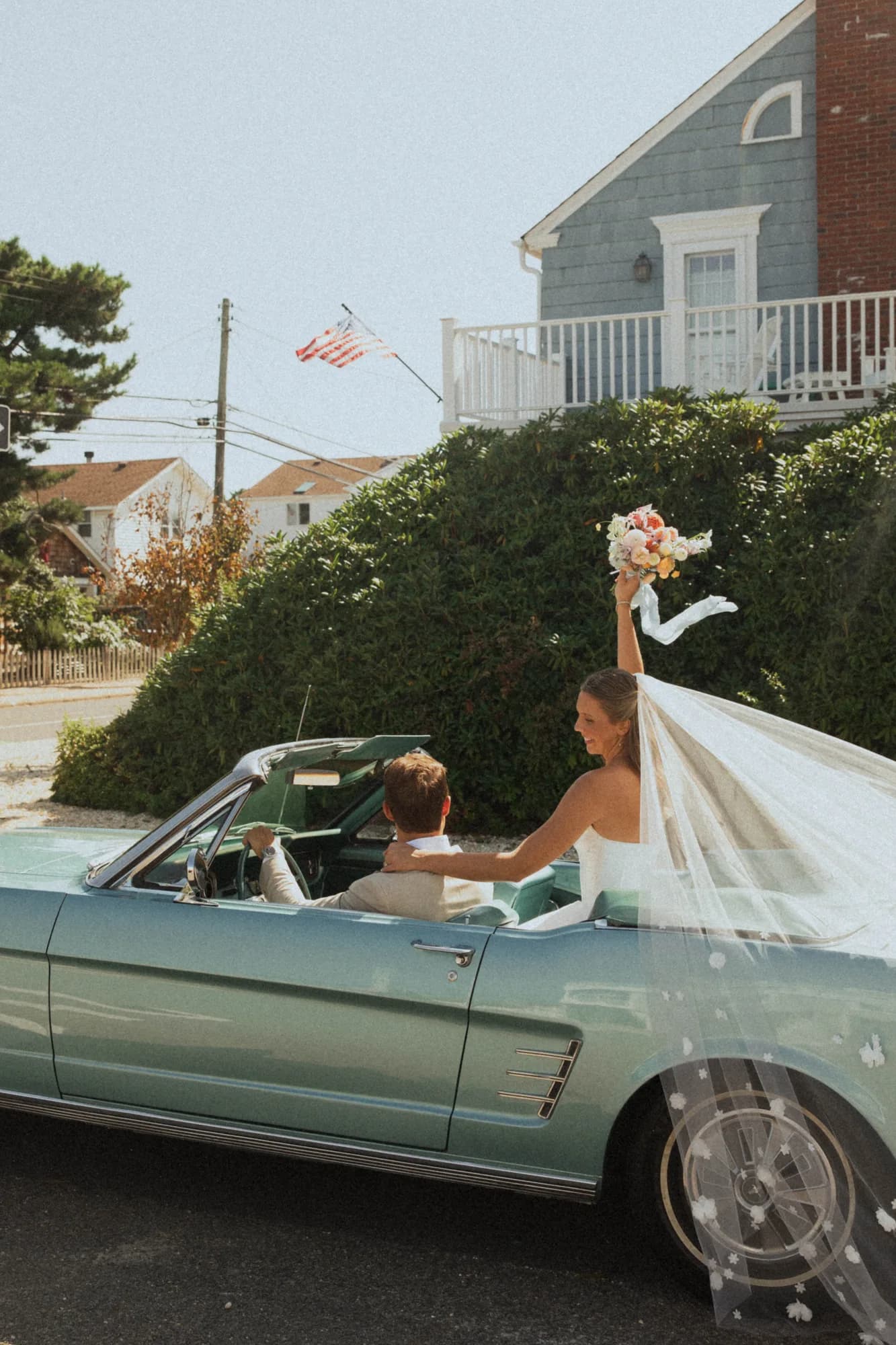 Talea raises her bouquet from the seat of a pale blue convertible, veil draped over the door, Erich at the wheel