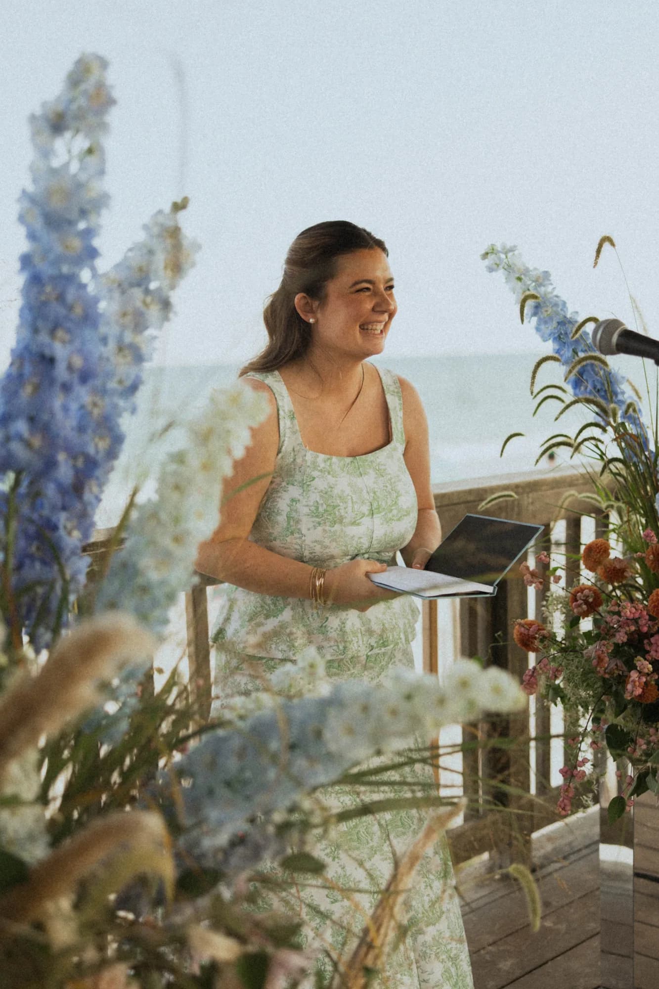 Officiant laughs at the altar framed by blue delphinium spires and grasses, orange dahlias and the sea beyond