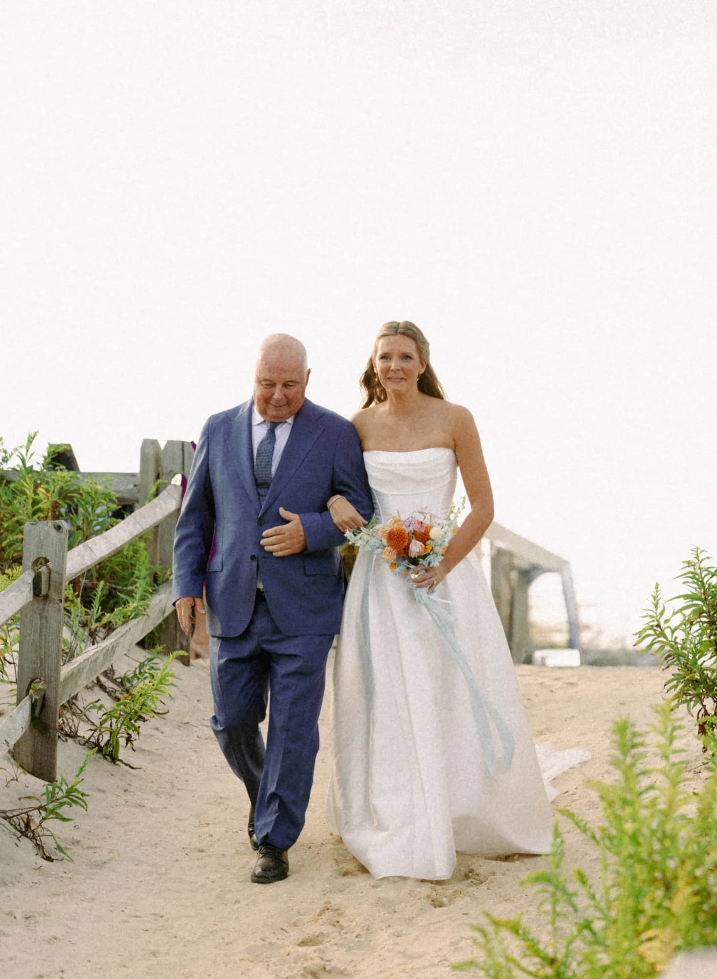 Talea walks arm in arm with her father along a sandy dune path, her bouquet trailing pale blue ribbon