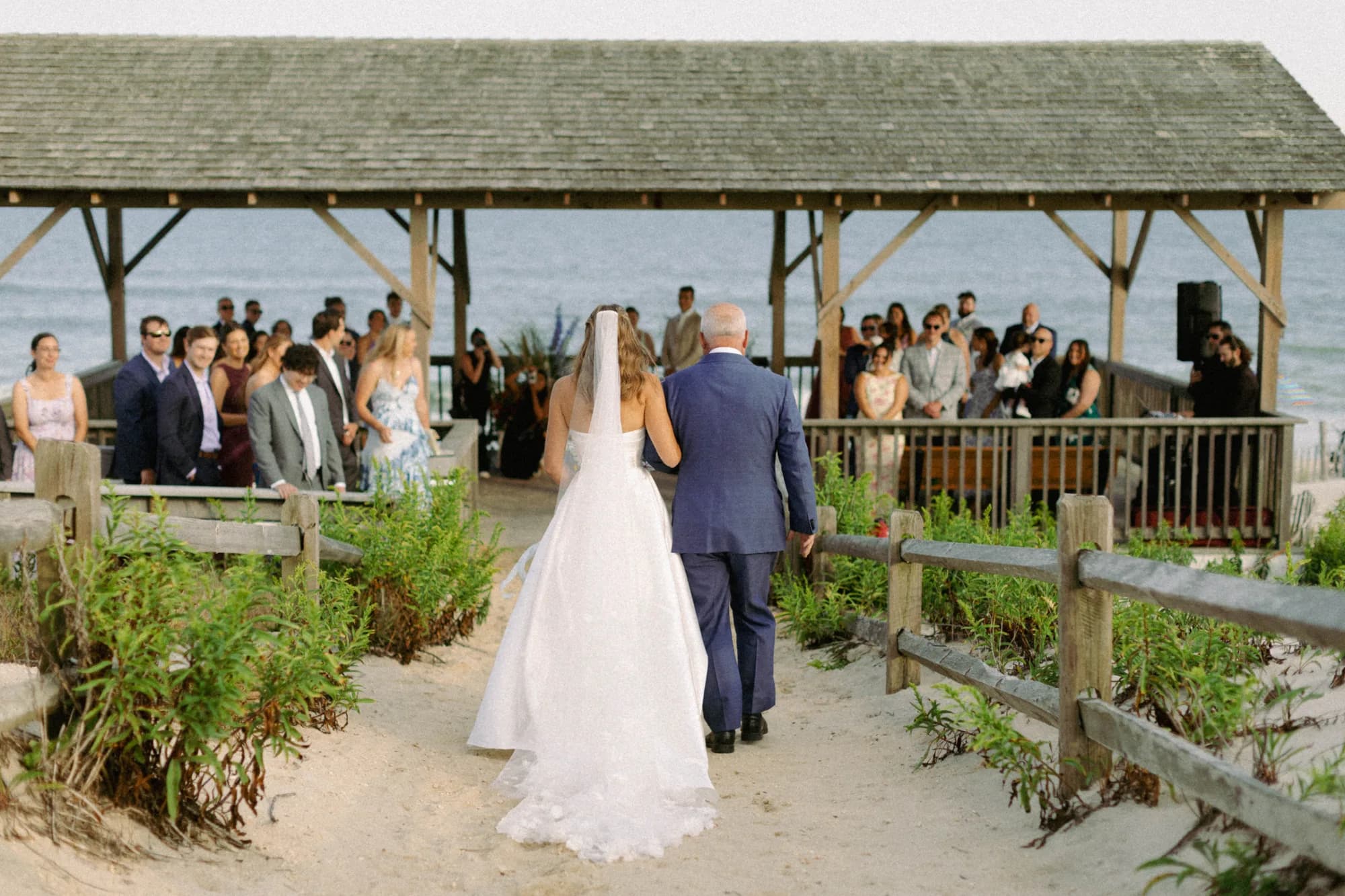 Talea and her father walk the sandy aisle toward the pavilion as guests stand, the ocean beyond