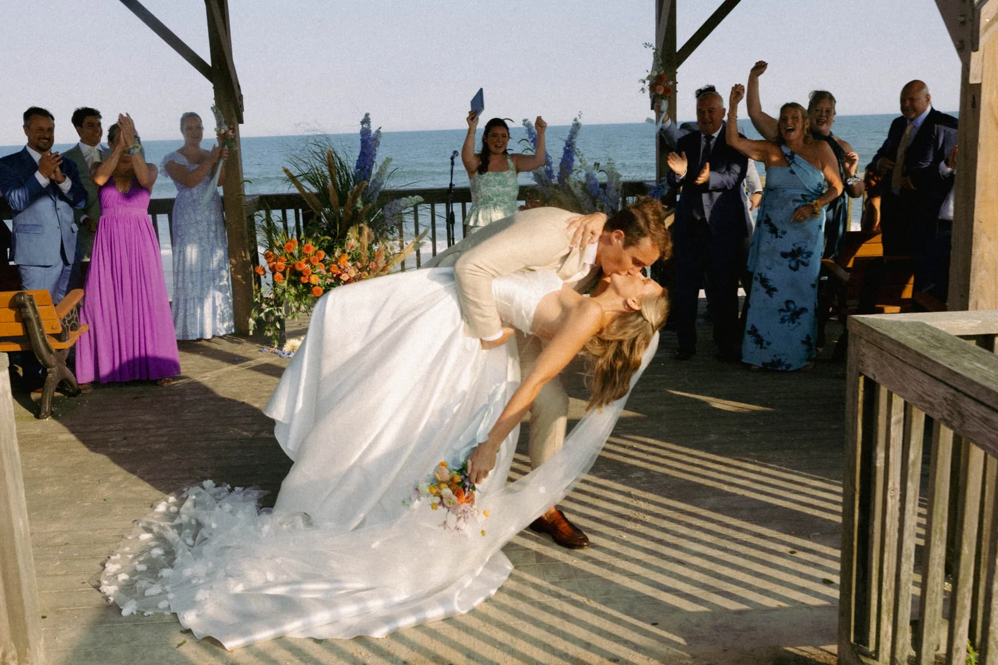 Erich dips Talea in a kiss on the pavilion deck as guests cheer, her bouquet in hand, arrangements behind
