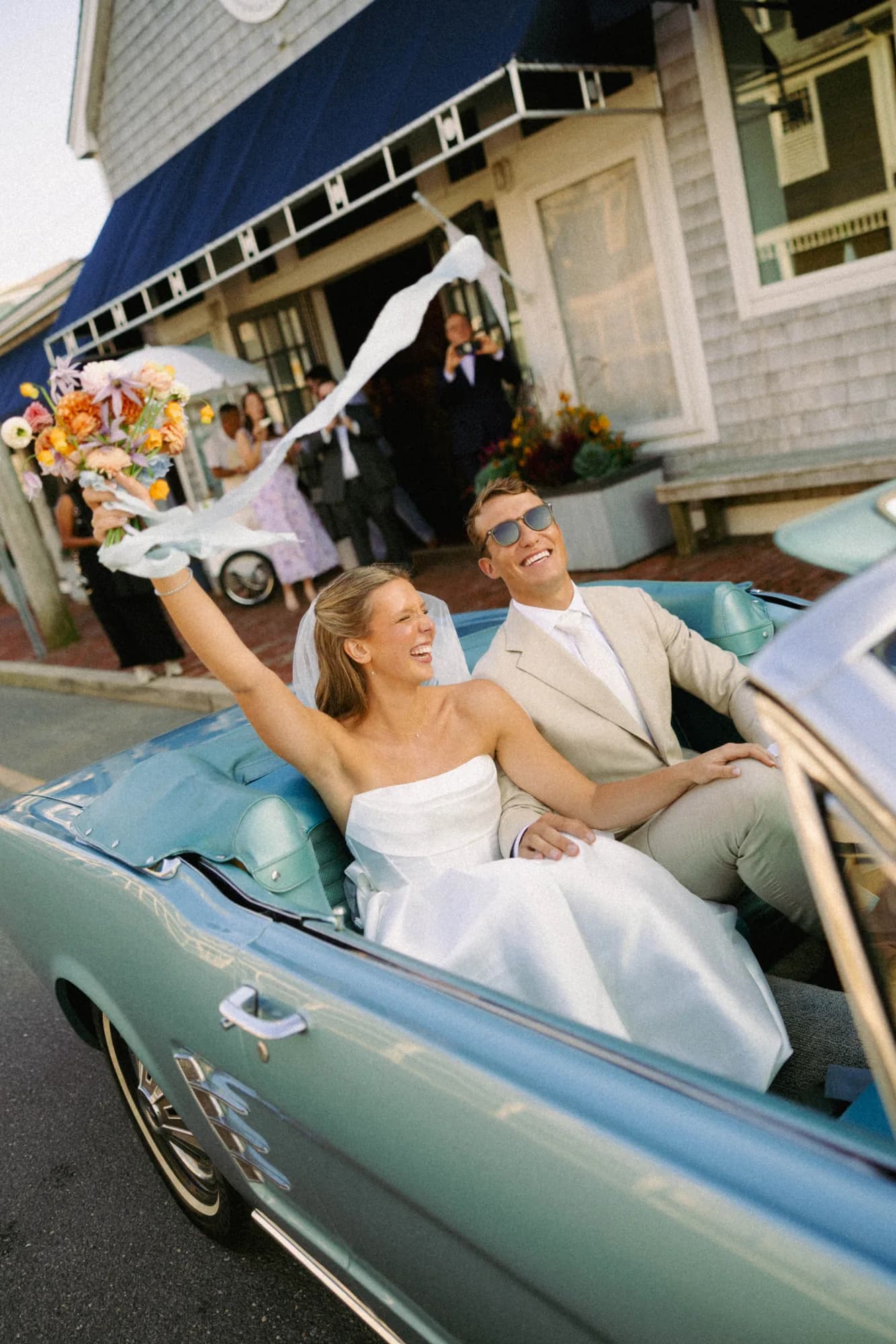 Talea raises her bouquet with trailing white ribbon from a pale blue convertible, Erich in sunglasses beside her