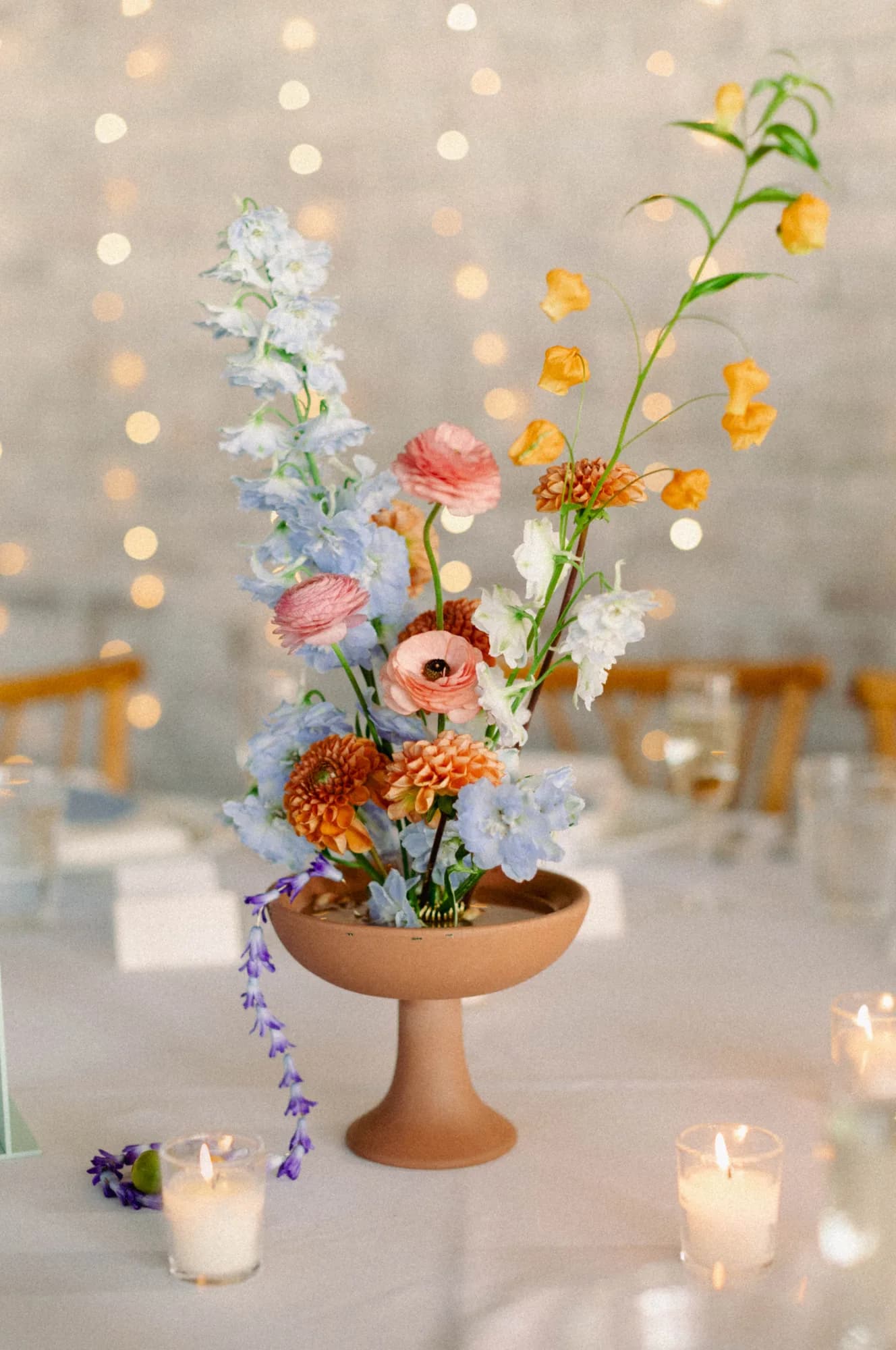 Terracotta compote of pale blue delphinium, pink ranunculus, orange dahlias and arching sandersonia, lights behind