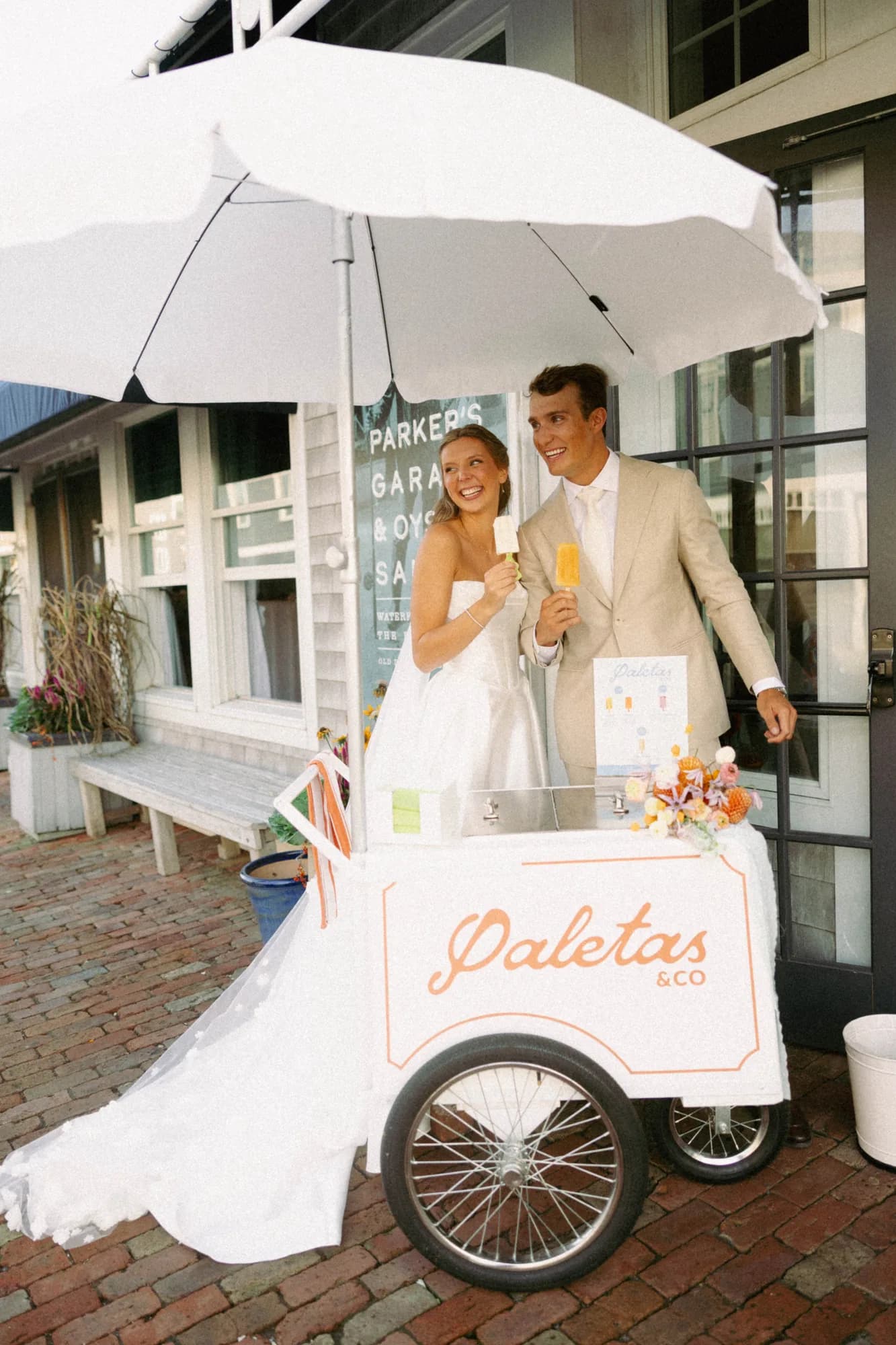 Talea and Erich eat ice pops beneath a white umbrella beside a cart dressed with orange dahlias and clematis