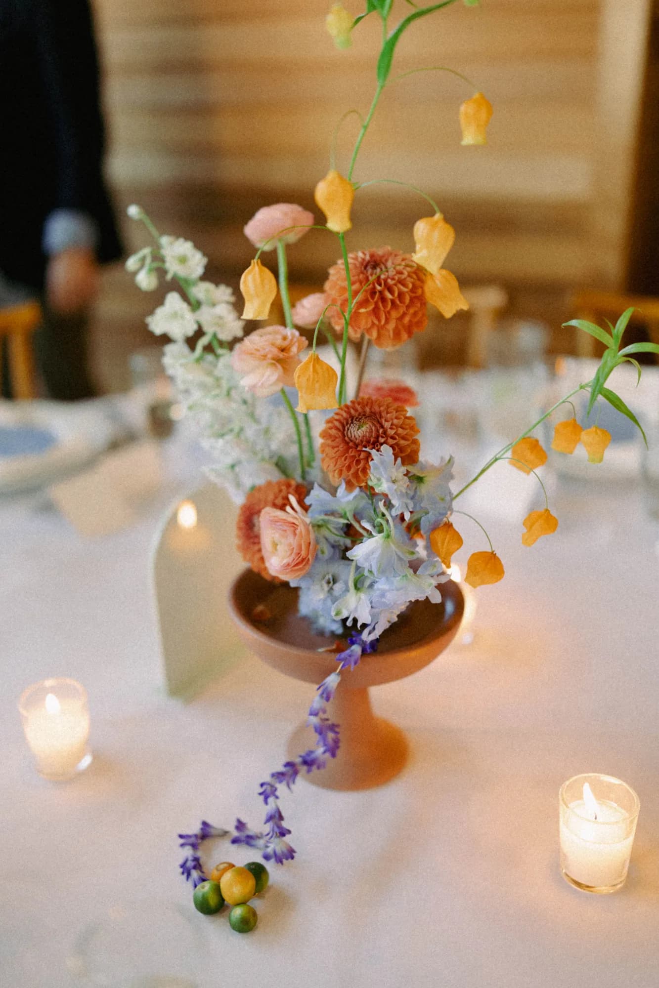 Terracotta compote from above with orange dahlias, peach ranunculus, blue delphinium and sandersonia lanterns