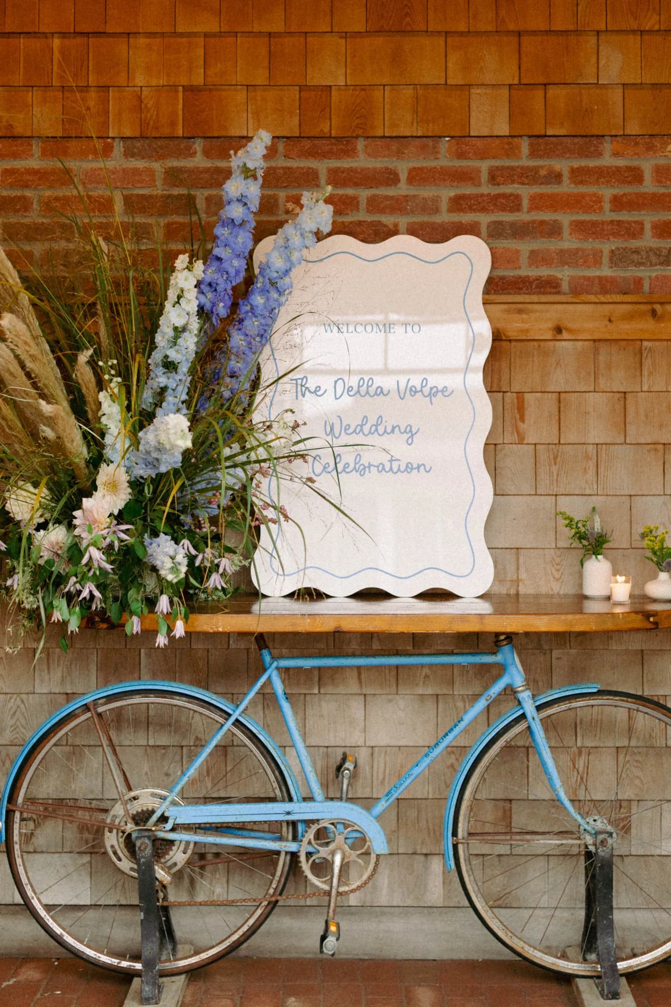 Welcome sign on a wooden table over a blue vintage bicycle, blue delphinium and dried grasses arranged beside