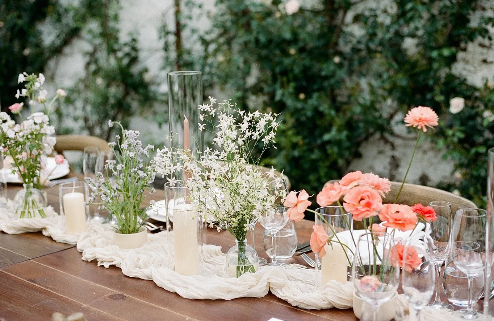 Clustered bud vases of coral ranunculus, pale blue amsonia and white lace flower run along a gauze runner between candles
