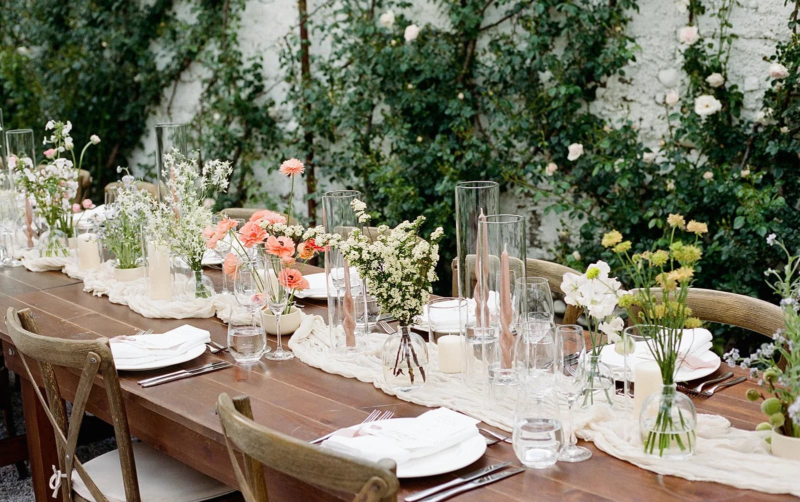 Long wooden reception table with bud vases of coral ranunculus, spirea and sweet pea, twisted blush tapers and roses behind