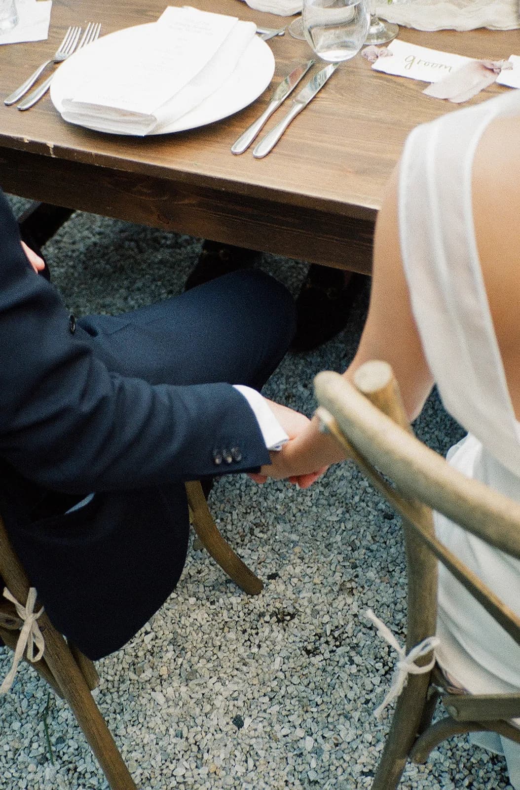 Trisha and Jim hold hands beneath the reception table, seated in crossback chairs on a gravel courtyard floor