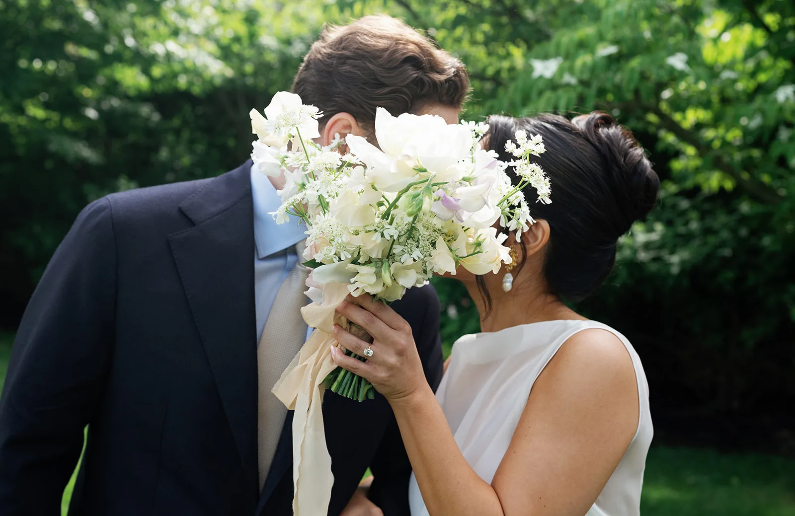 Trisha lifts her bouquet of white peonies and sweet pea to shield a kiss with Jim, silk ribbon trailing from the stems