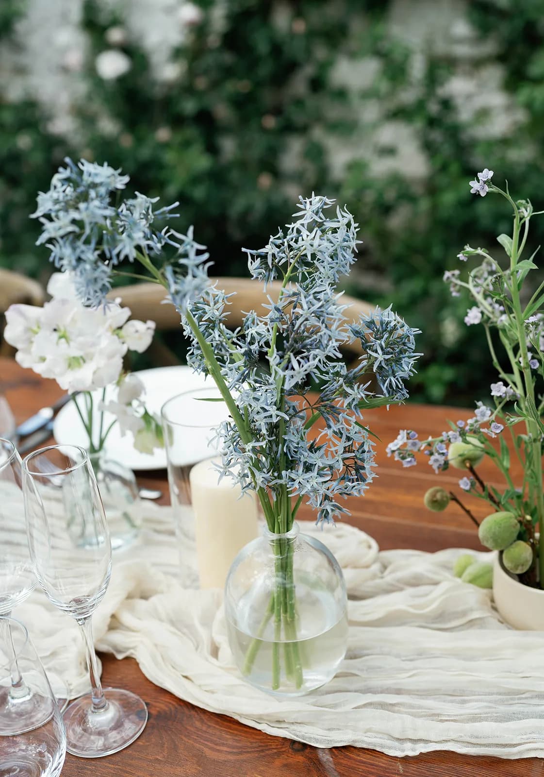 Pale blue amsonia in a round glass bud vase beside a pillar candle, with white sweet pea and a bowl of baby peaches behind