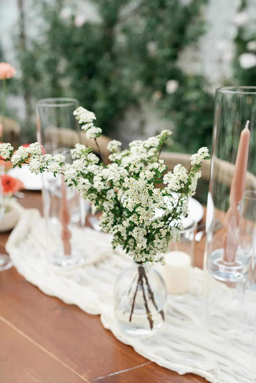 Spirea branches arch from a round glass bud vase on a gauze runner, twisted blush tapers standing in glass hurricanes behind