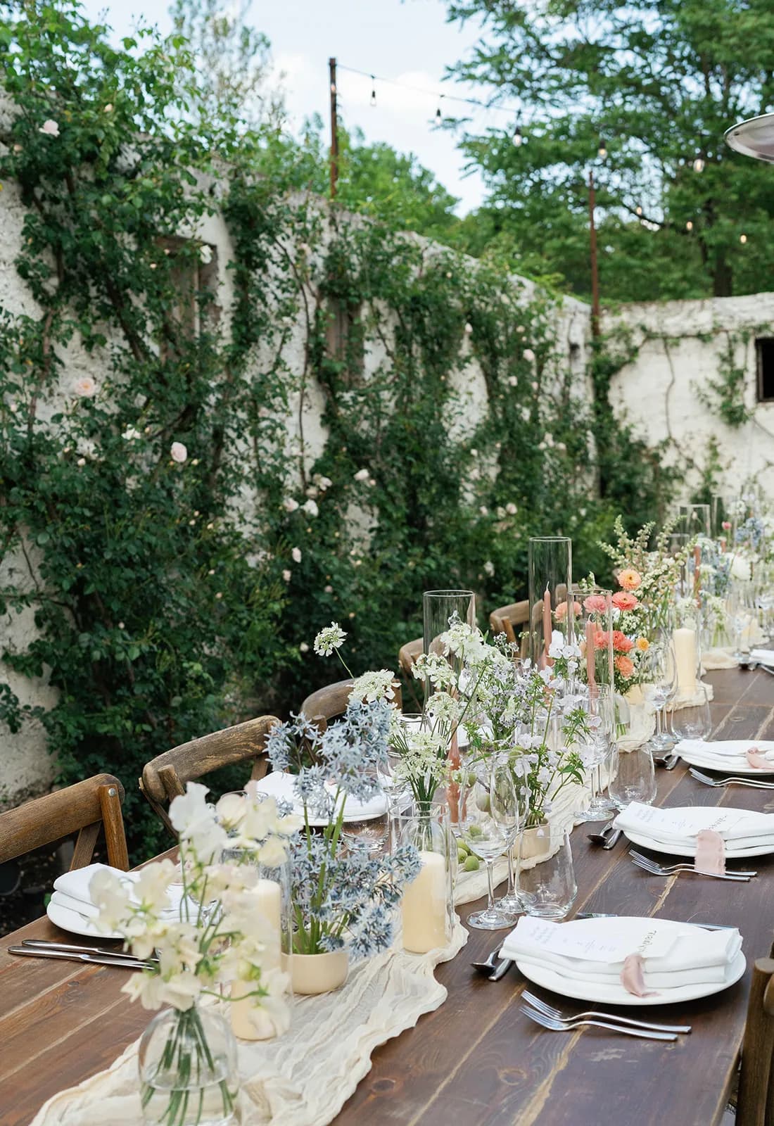 The long reception table seen from its end, bud vases of sweet pea, amsonia and coral ranunculus threading between candles