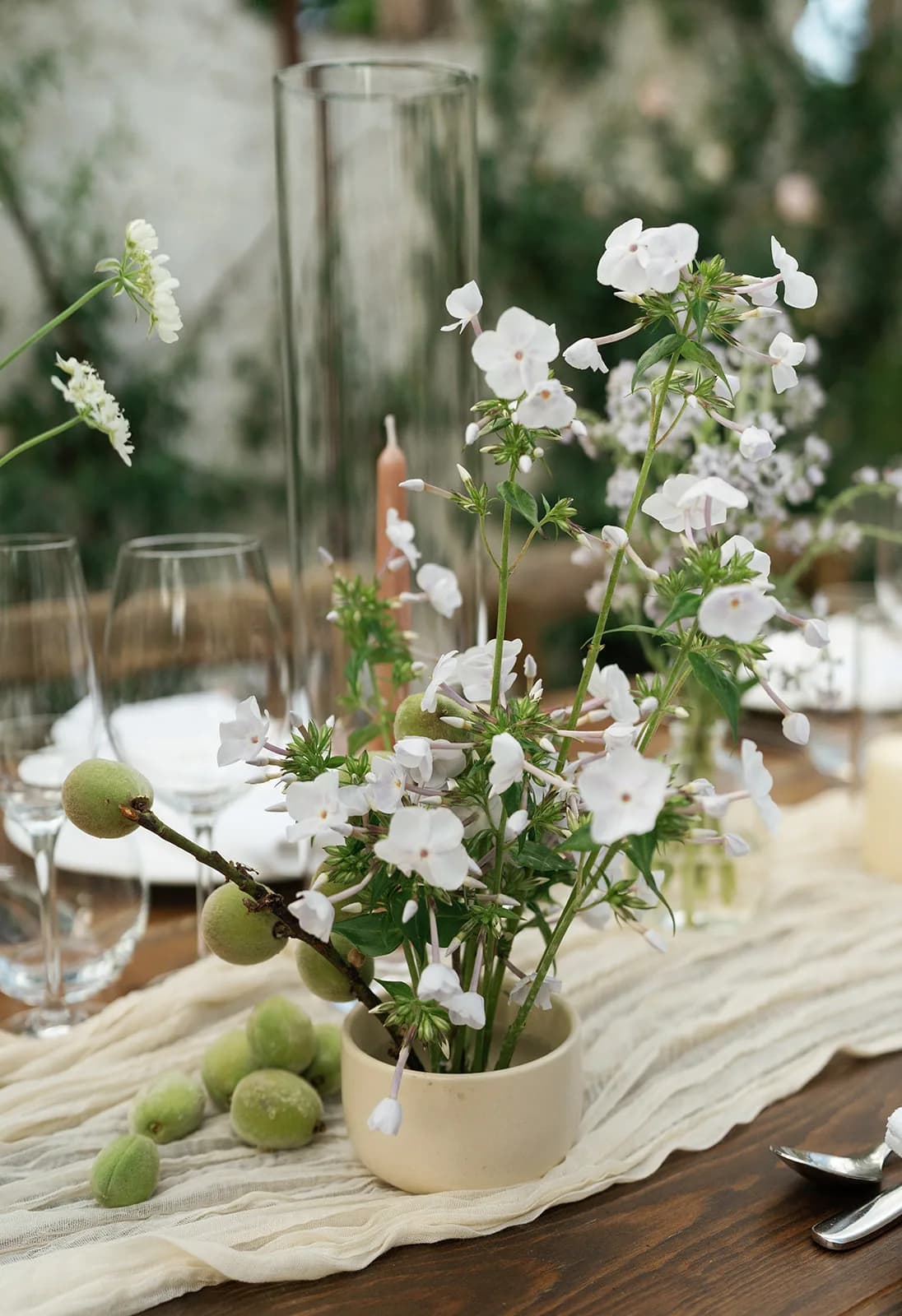White phlox rises from a low cream ceramic bowl as a baby peach branch rests across it, loose peaches on the gauze runner