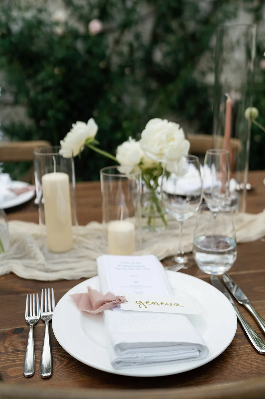 A guest's place setting with menu and blush silk ribbon, white peonies in bud vases and pillar candles in glass behind
