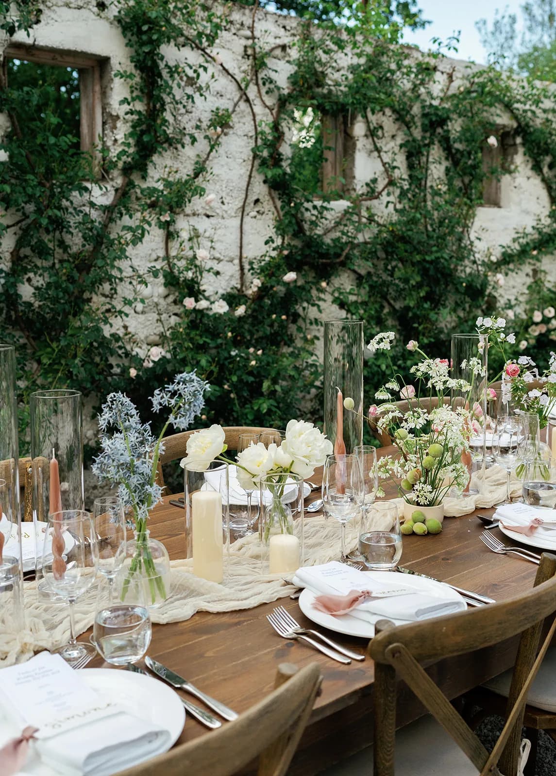 Tablescape of pale blue amsonia, white peonies, lace flower and baby peaches below a whitewashed wall of climbing roses