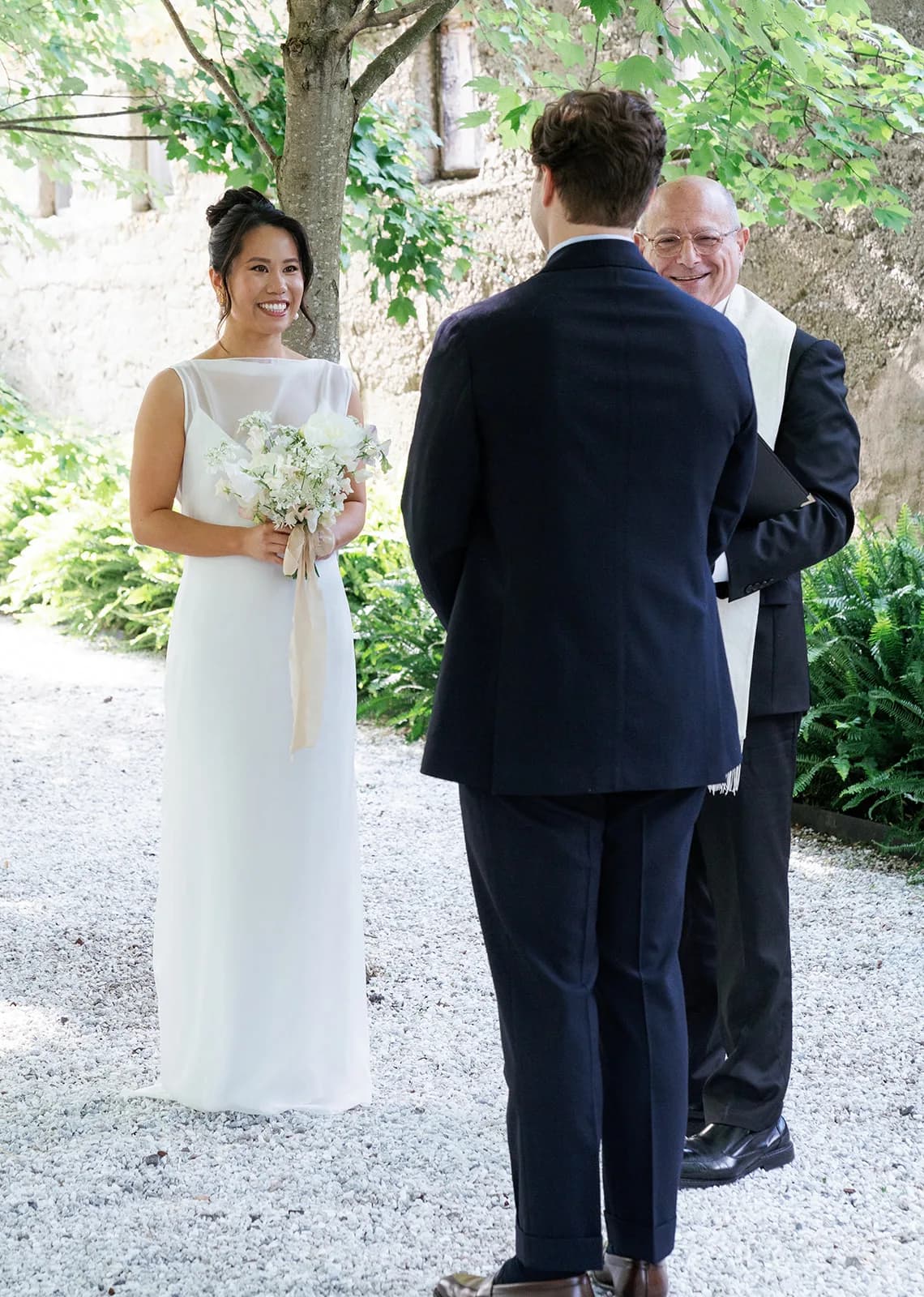 Trisha smiles at Jim during the ceremony, holding her petite bouquet of white peonies and sweet pea with a long silk ribbon